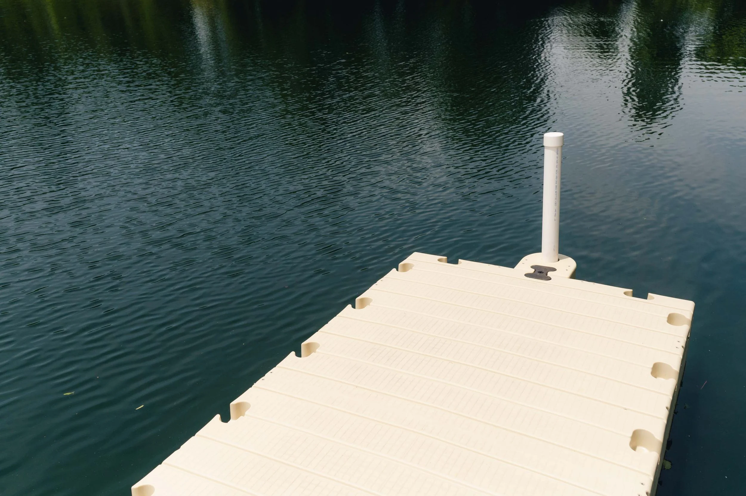 A beige floating dock with a white pole extends into a calm body of water, surrounded by trees and sky reflections.