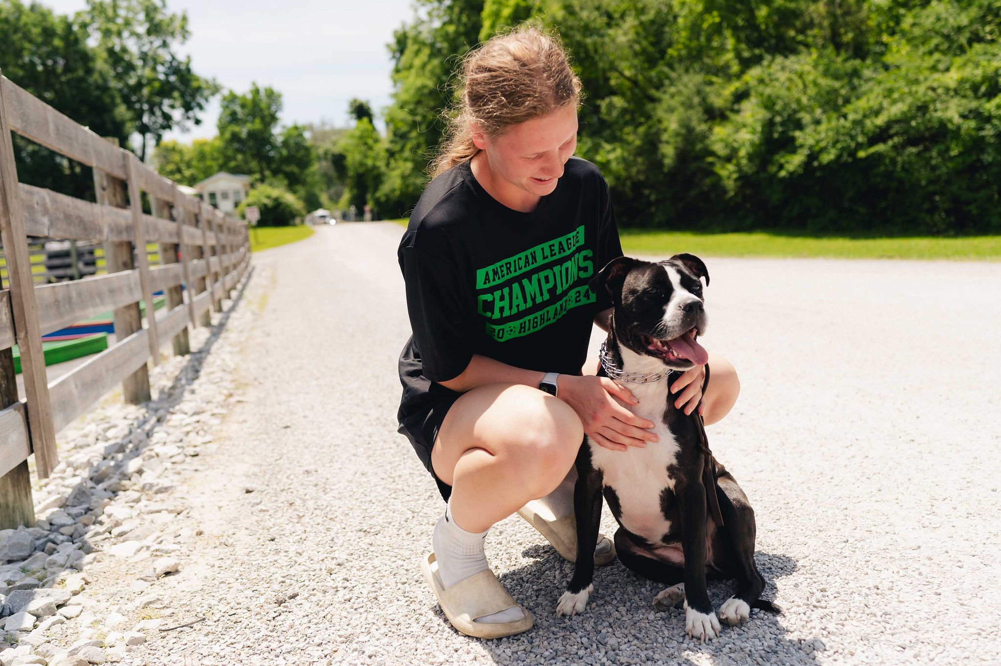 A young woman in a black t-shirt and shorts is kneeling on a gravel path outdoors, smiling and holding a black and white dog with its tongue out. The scene is sunny with green trees in the background.