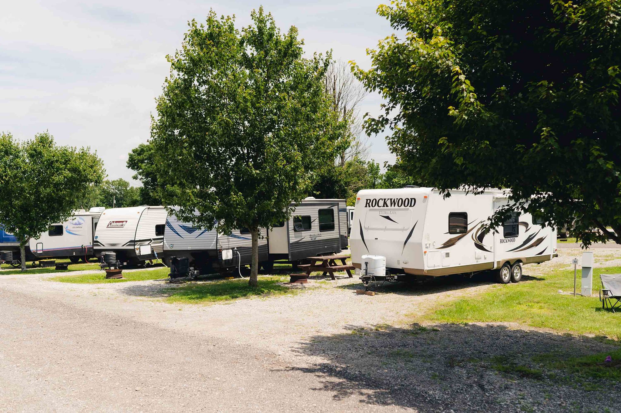 A row of recreational vehicles (RVs) parked at a campsite with green trees and a gravel pathway.
