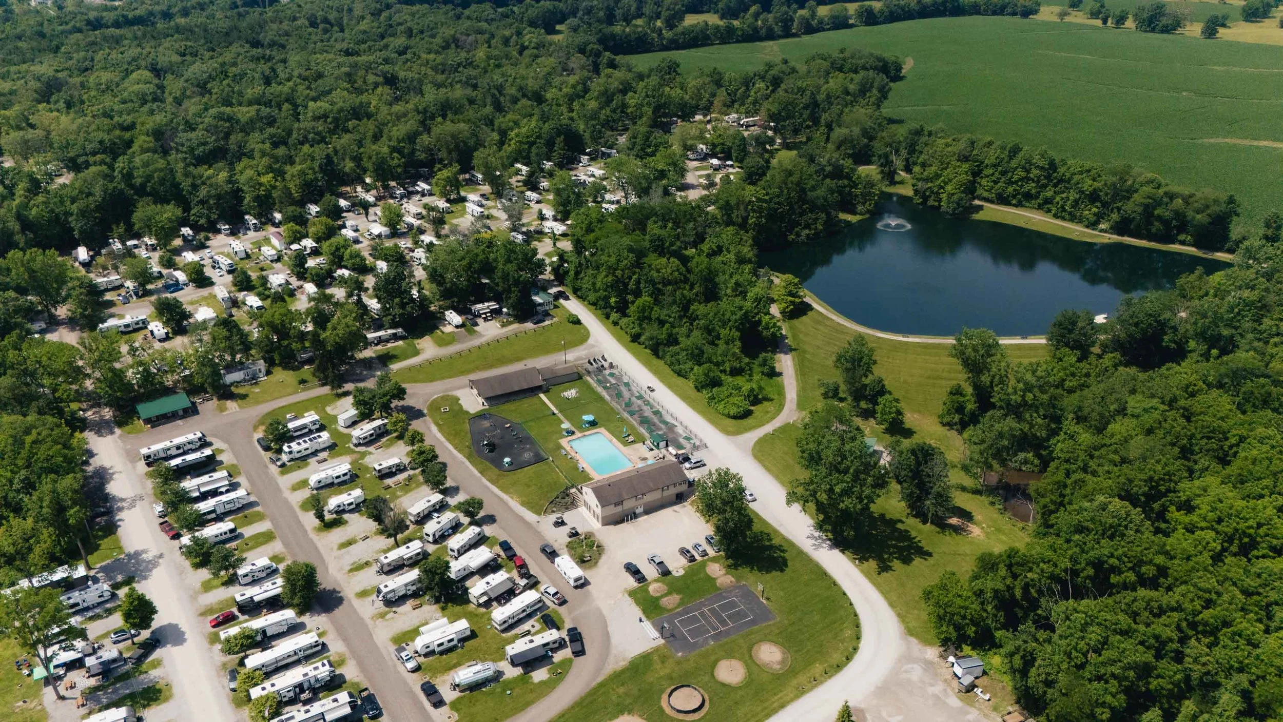 Aerial view of a campground with numerous RVs and camper trailers, a swimming pool, a pond with a fountain, and surrounding green trees and grass.