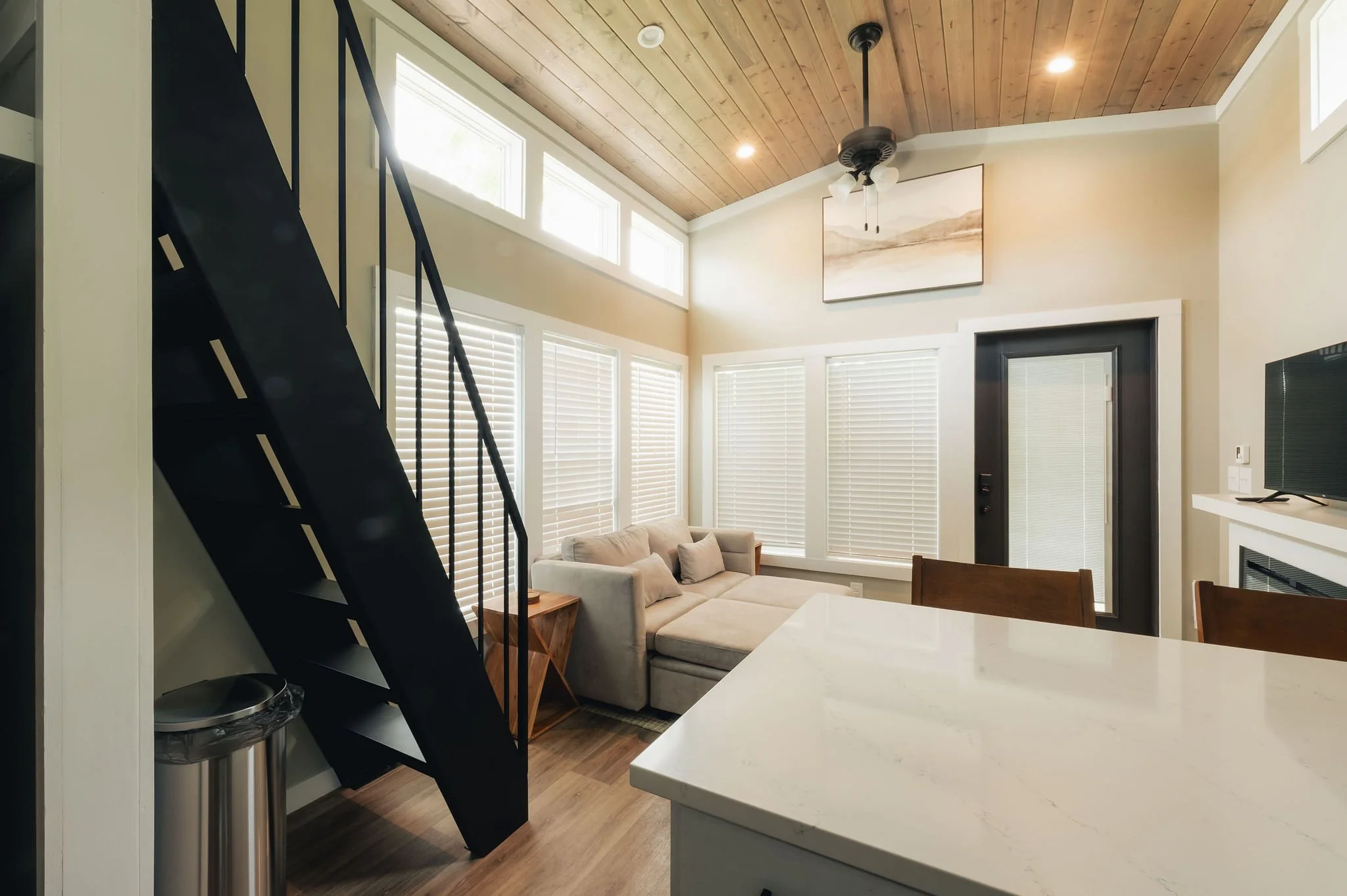 Living room with beige sectional sofa, wooden side table, large windows with white blinds, black front door, and a wooden ceiling with a ceiling fan