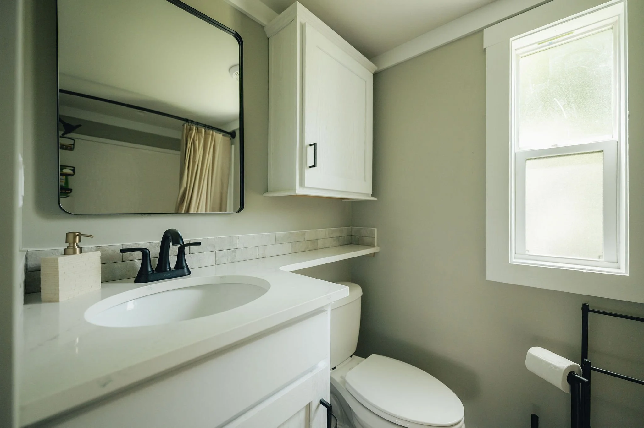 Small bathroom with white vanity, black faucet, mirror, closed cabinet, and a window with frosted glass