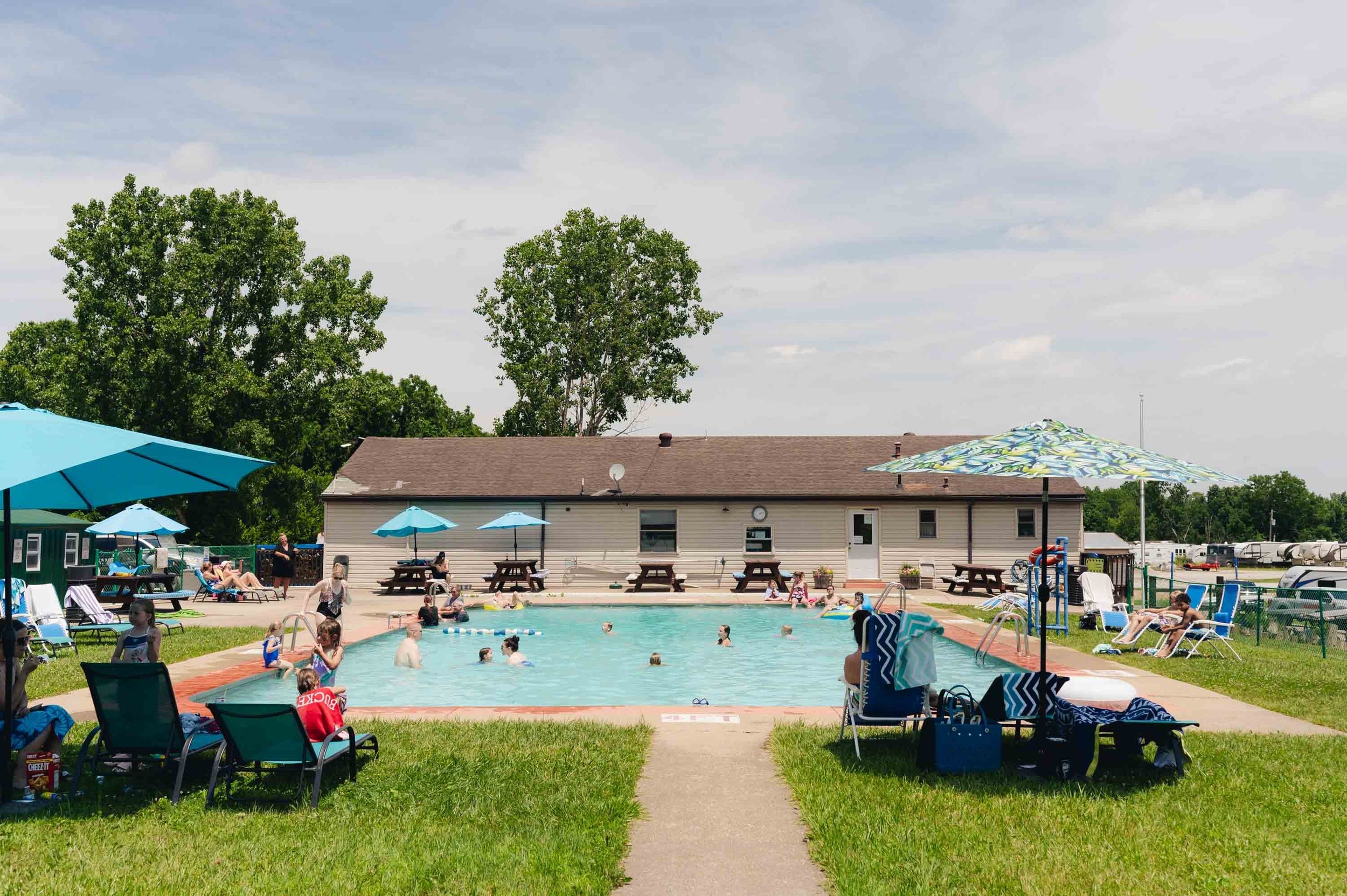 People enjoying swimming and relaxing at a public pool with umbrellas and lounge chairs, during a sunny day.