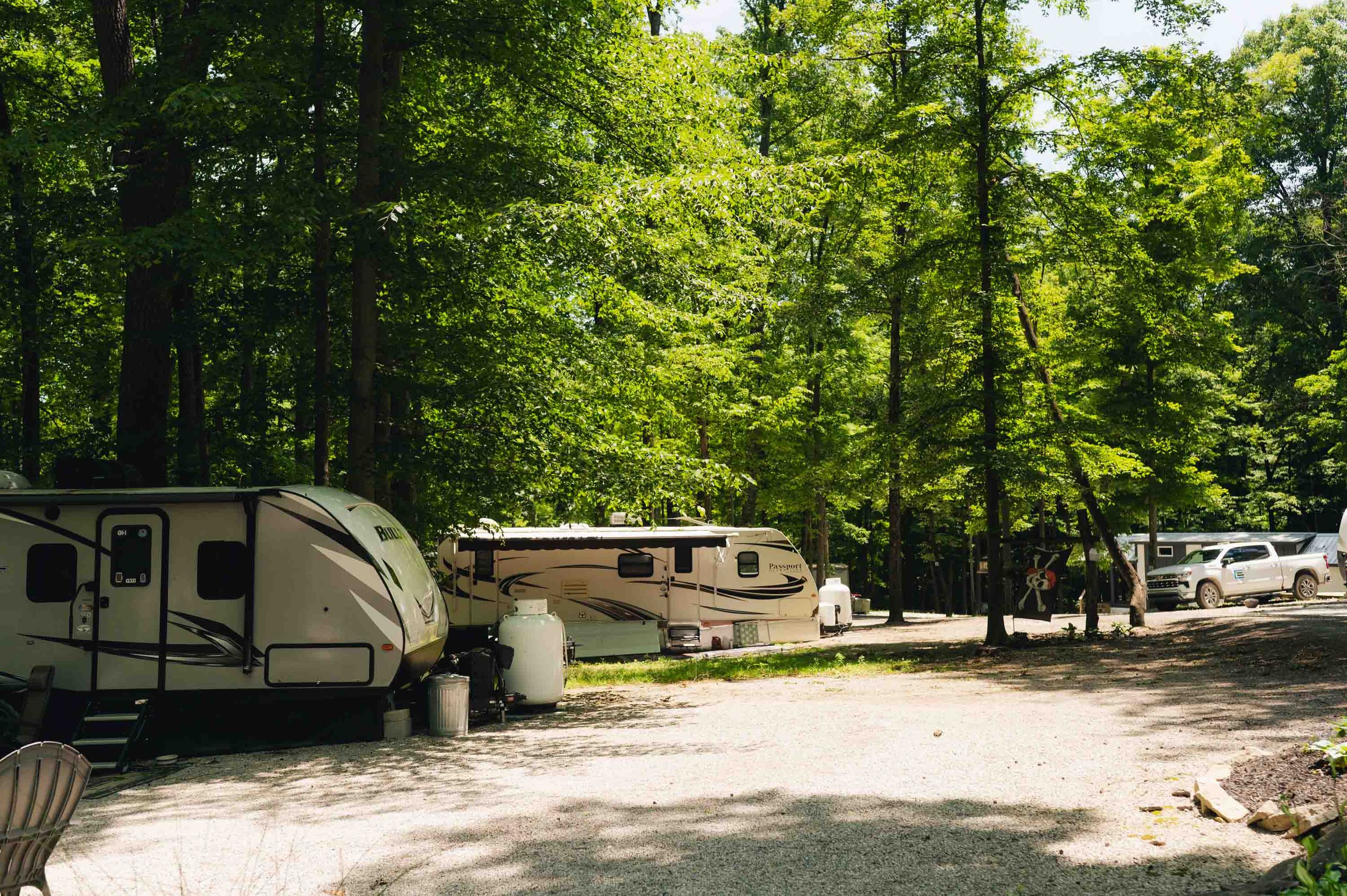 Campground with trees, two parked RV trailers, outdoor chairs, and several vehicles in the background.