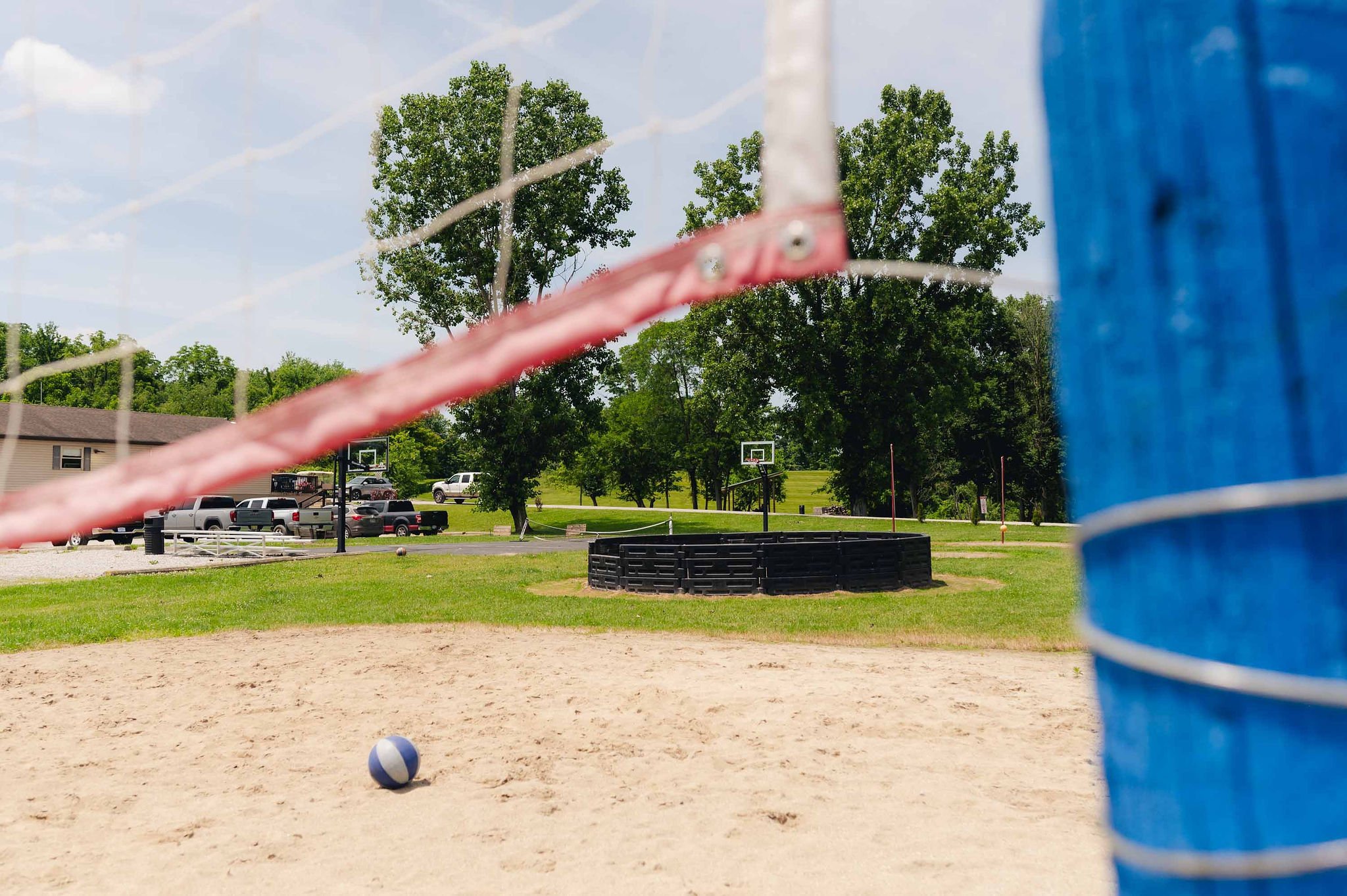View through a volleyball net of a sandy volleyball court with a volleyball on the sand, a grassy area with trees, basketball hoops, parked cars, and a racetrack in the background.