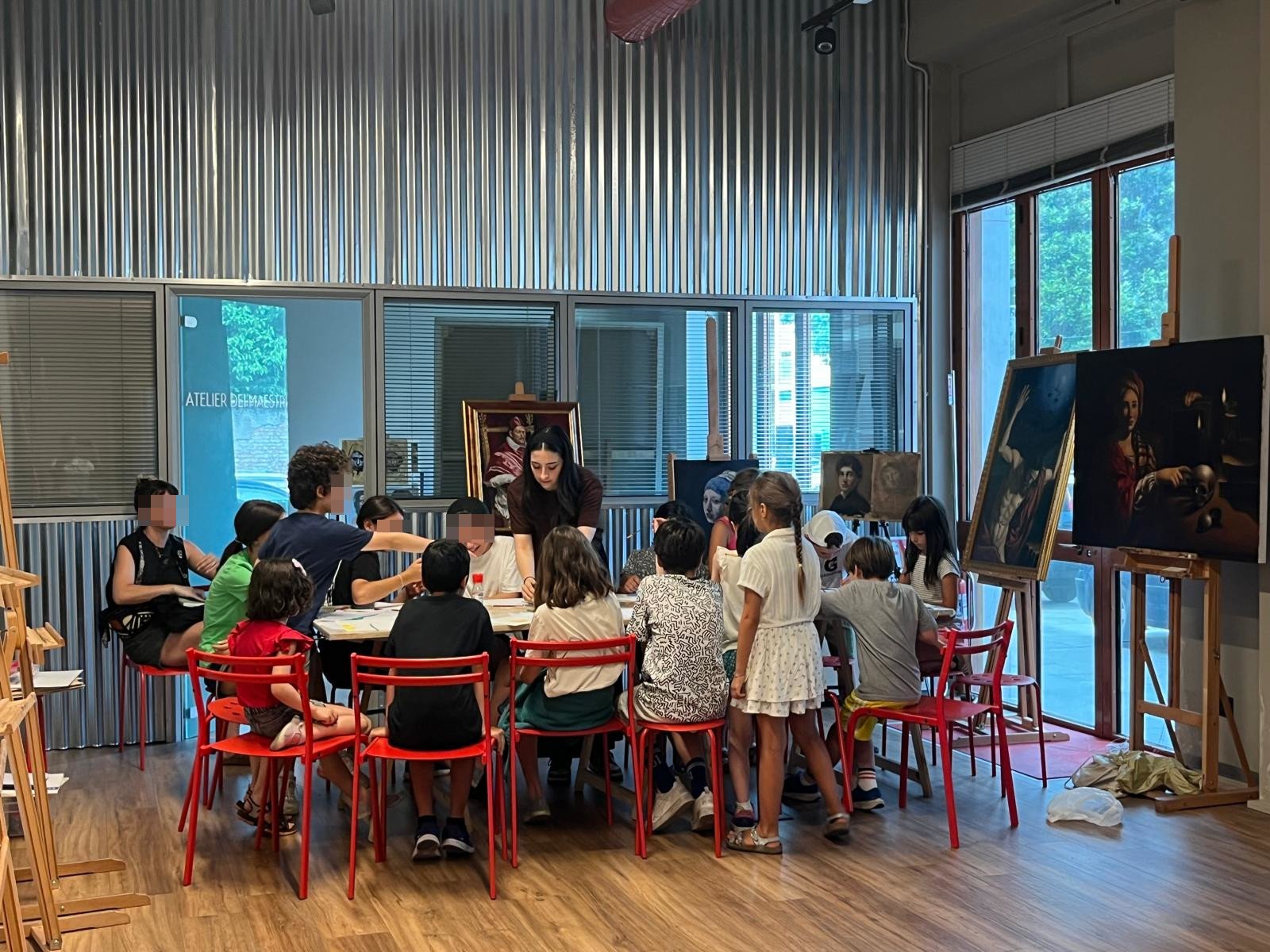 Corso di disegno e pittura per bambini seduti intorno a un tavolo in un atelier d'arte, mentre un adulto li assiste. La stanza ha grandi finestre con luce naturale e dipinti appesi alle pareti e su cavalletti.