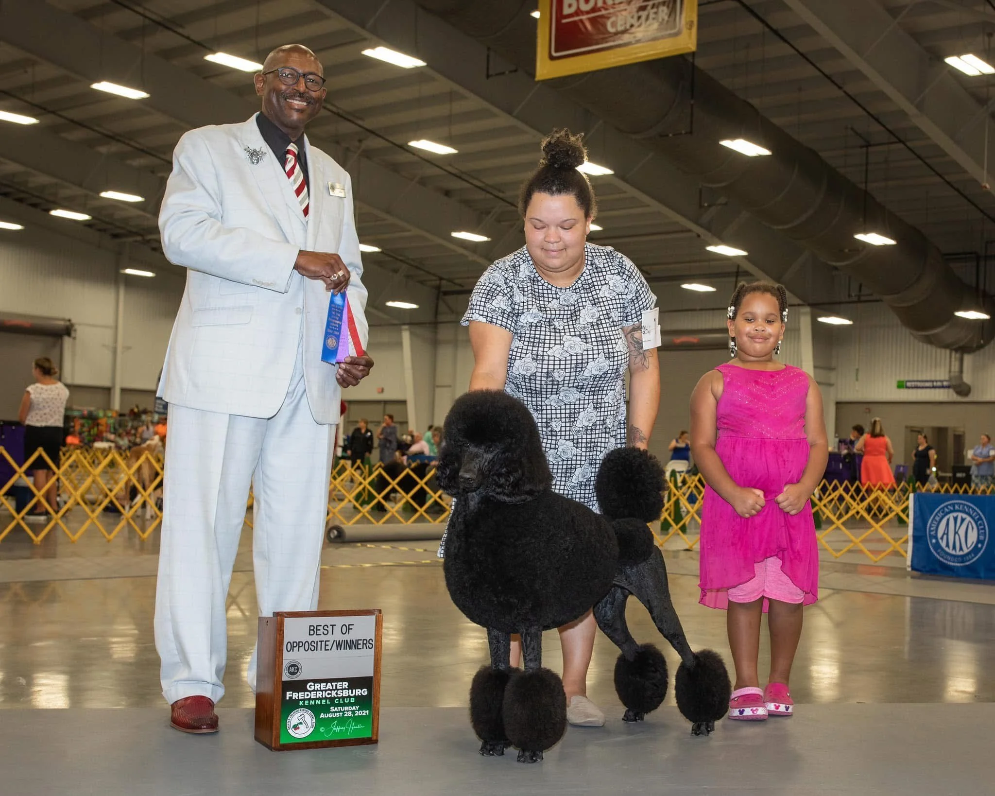 A man in a white suit holding a ribbon, a woman with a poodle, and a girl in a pink dress standing inside an indoor dog show arena with a sign that says 'Best of Opposite/Winners'.