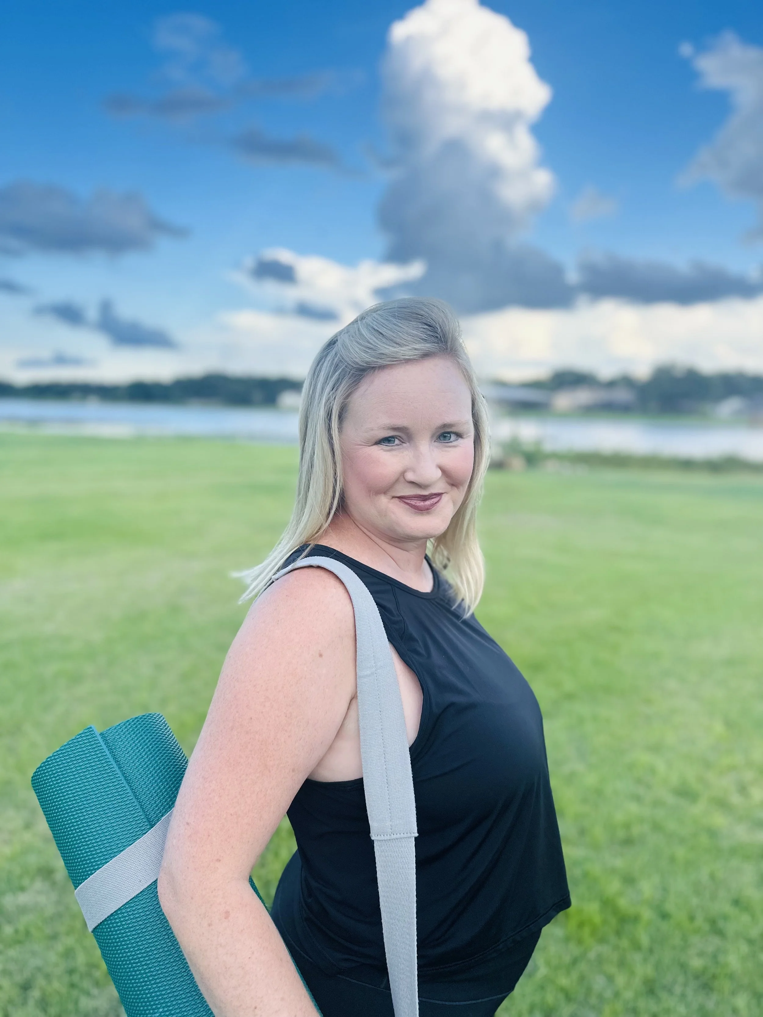 A woman with blonde hair, dressed in a black sleeveless top, outdoors on a grassy field with water and a cloudy sky in the background, carrying a blue and gray yoga mat on her shoulder.