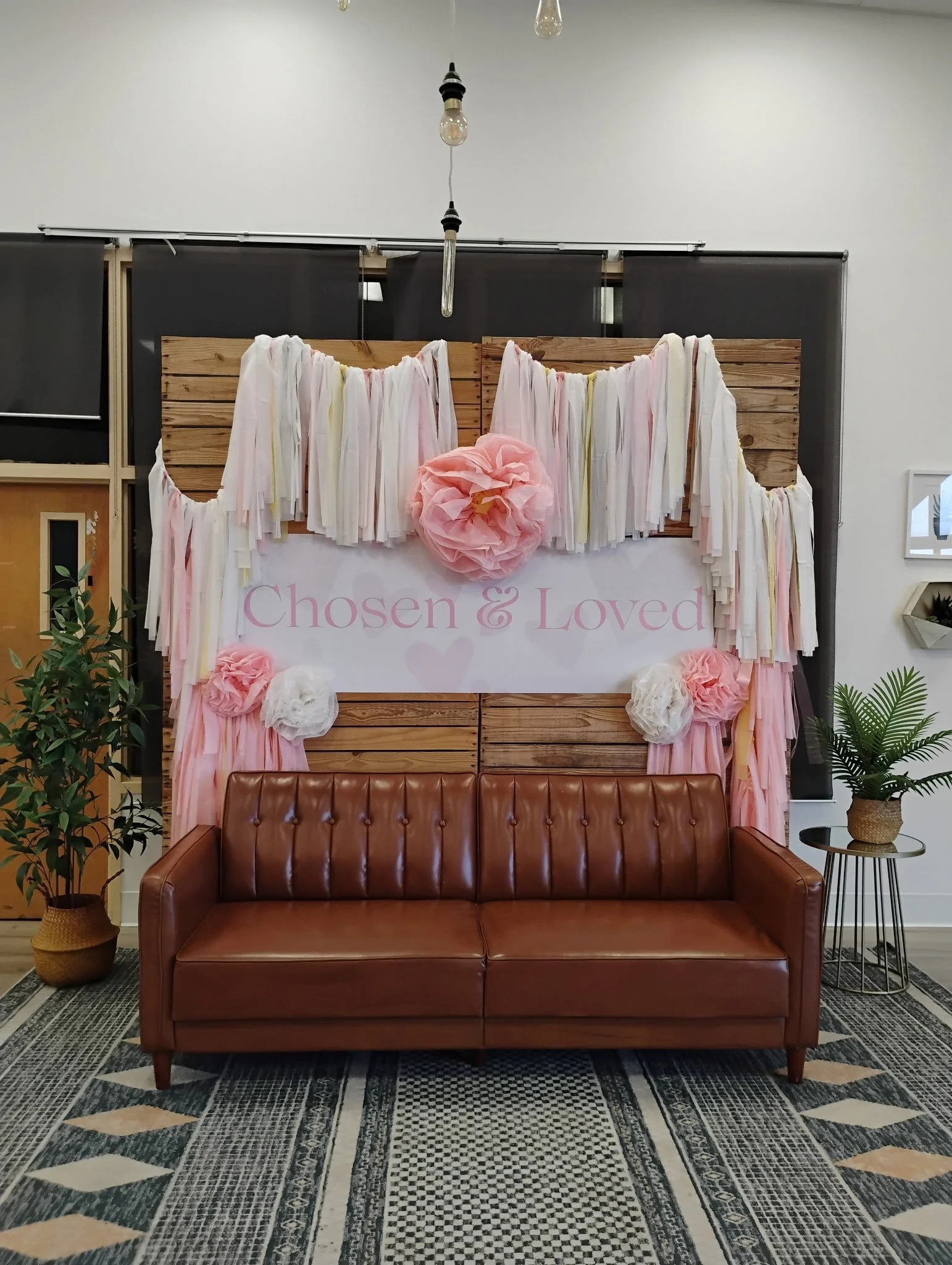 Decorative backdrop with pink and white tissue paper flowers, pastel-colored tassels, and a sign reading 'Chosen & Loved' in pink lettering, set up behind a brown leather sofa, with potted plants on either side.