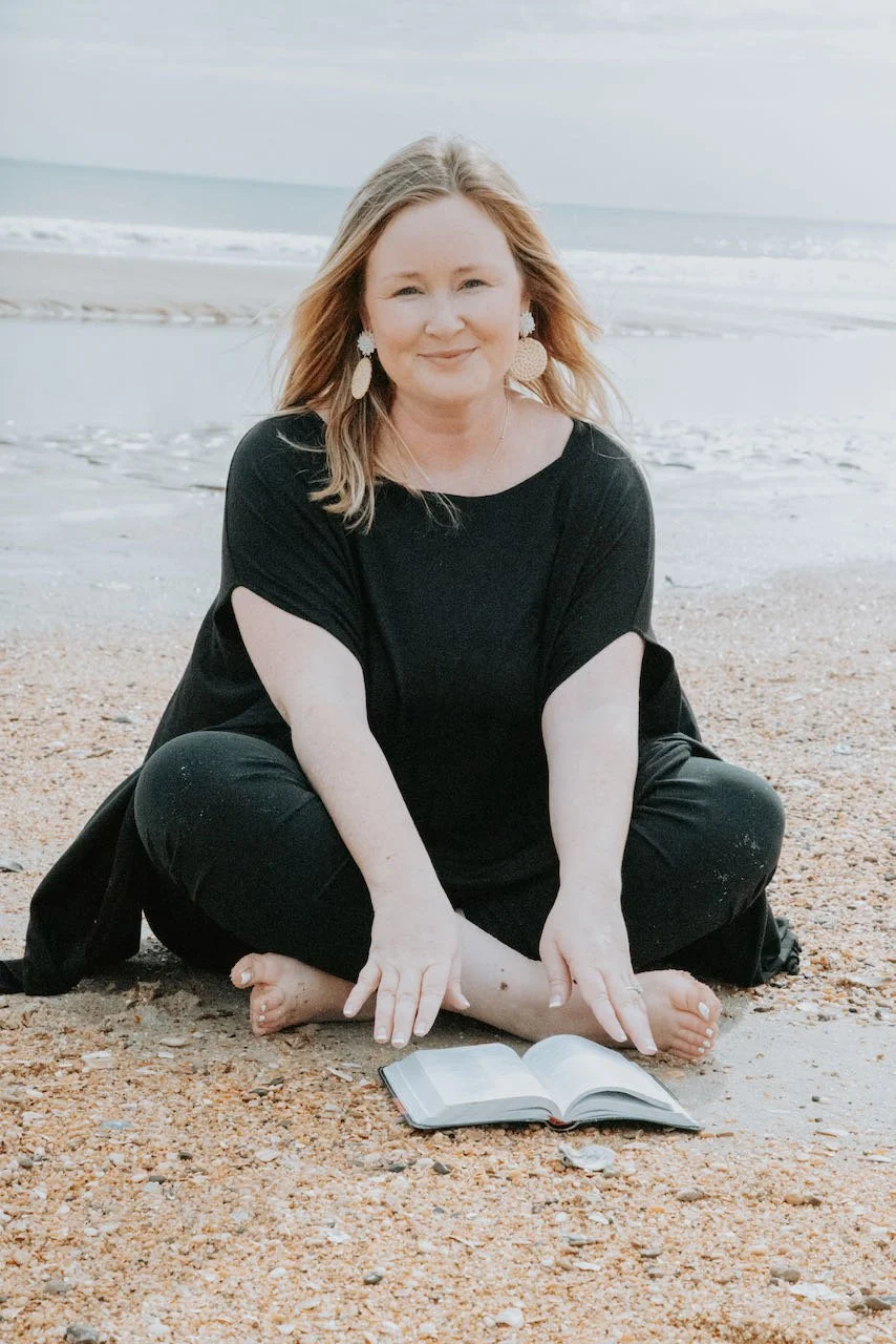 A woman with long blonde hair sitting cross-legged on a sandy beach, smiling, with an open book in front of her.