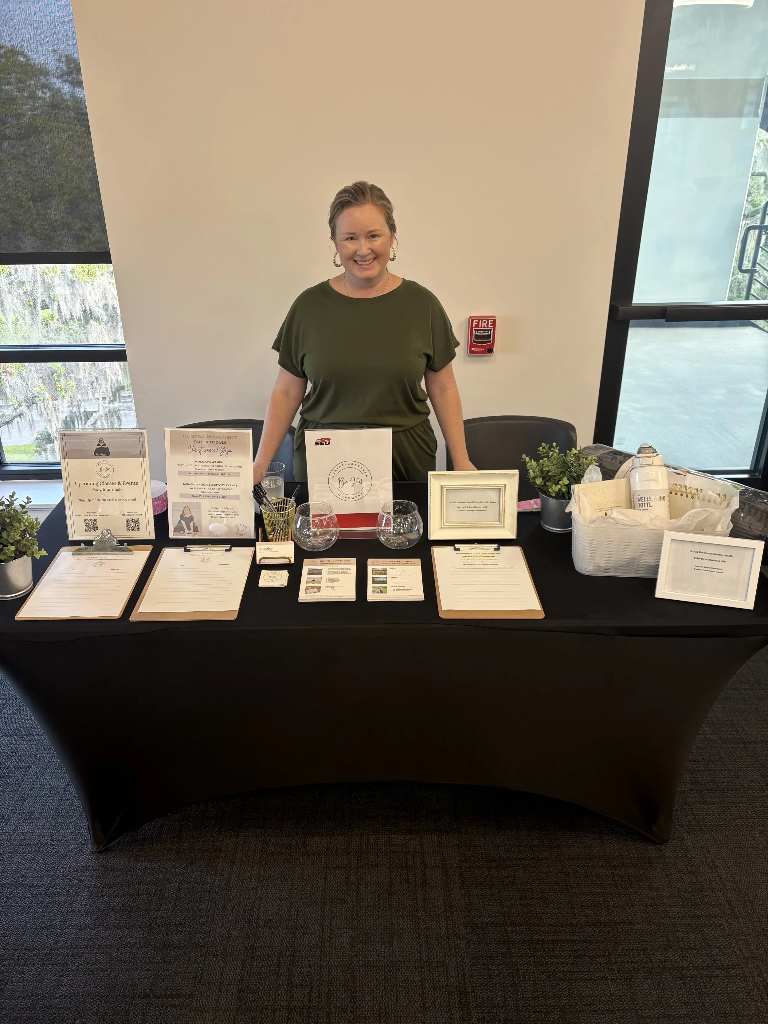 Woman standing behind a table at a registration or info booth in a modern building with large windows.