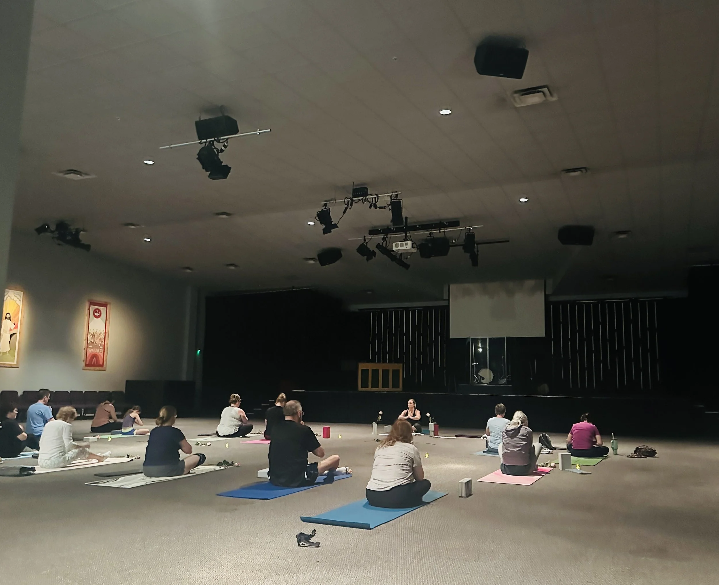 People participating in a yoga class in a dark room, sitting on yoga mats in a semi-circle facing a instructor at the front.