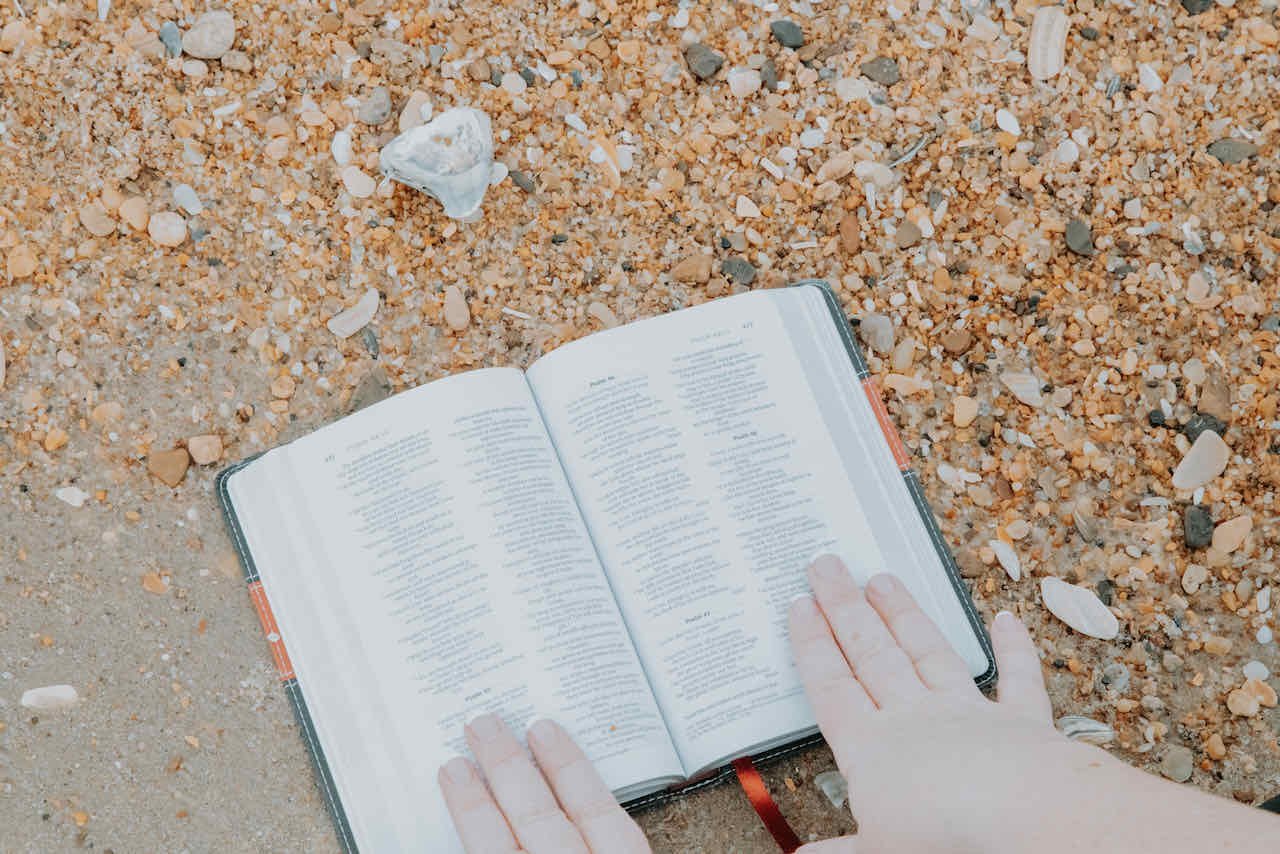 A person holding an open Bible on a sandy surface with small seashells scattered around.