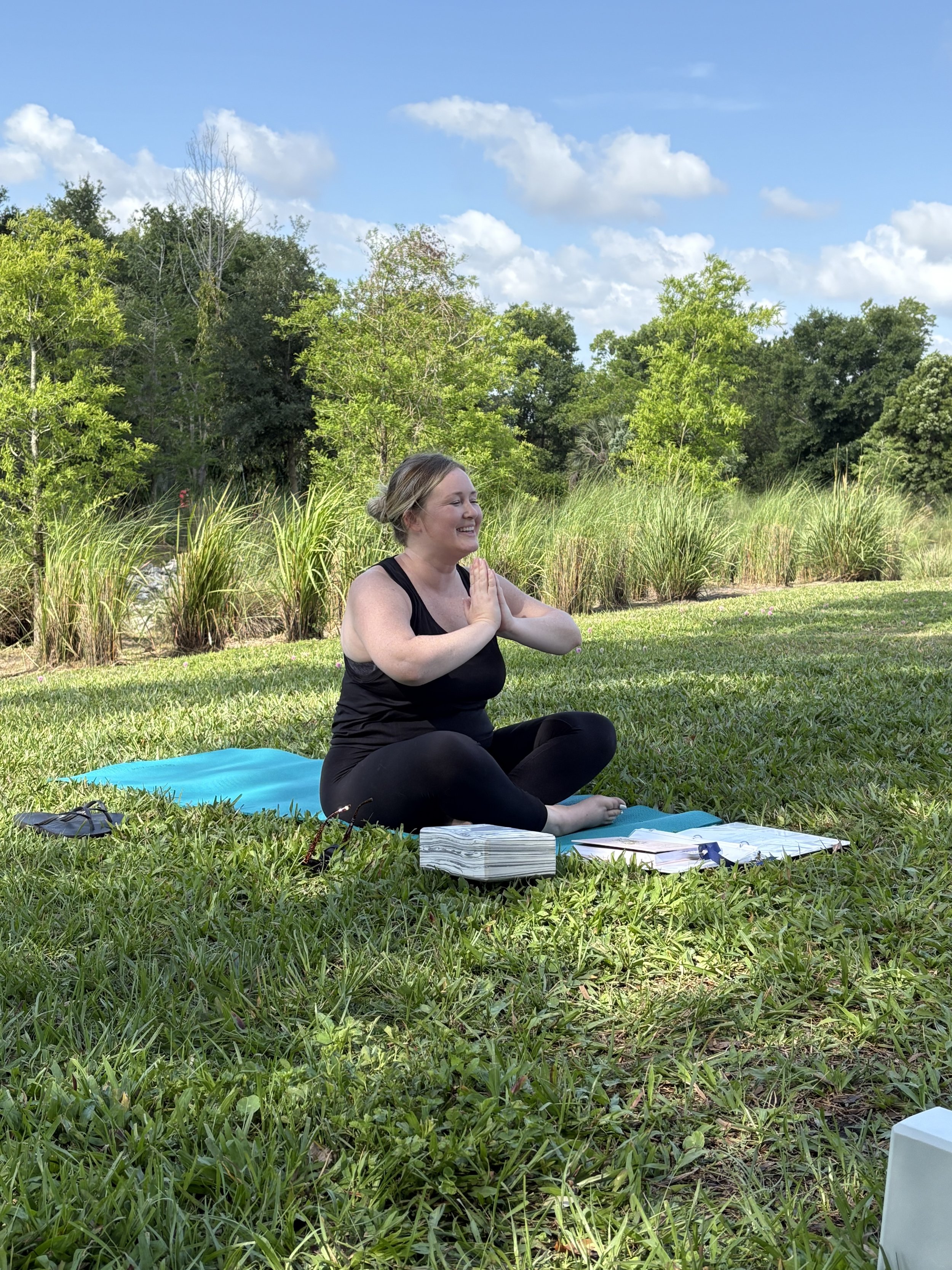 A woman practicing yoga outdoors on a blue mat in a grassy area surrounded by trees and bushes, with a partly cloudy sky.