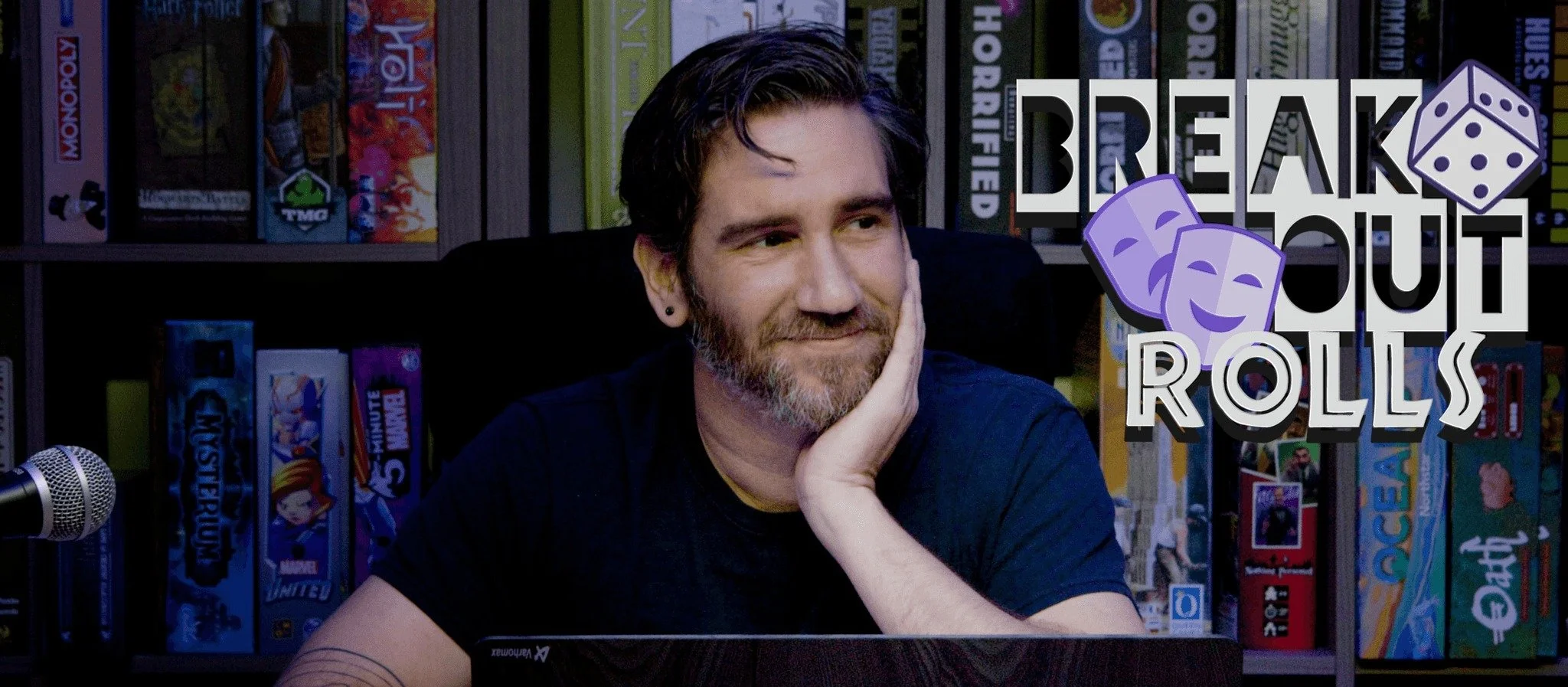 A man with a beard and dark hair, sitting at a table with his chin resting on his hand, in front of a bookshelf filled with board games, with a logo in the upper right corner that reads 'Breakout Rolls' and features two purple comedy masks and a purp
