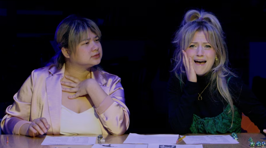 Two women sitting at a table with papers and poker chips. The woman on the left with short light brown hair, wearing a shiny pink jacket, appears concerned. The woman on the right with long blonde hair, wearing a black top with green patterns, looks distressed and is holding her face with her hand.