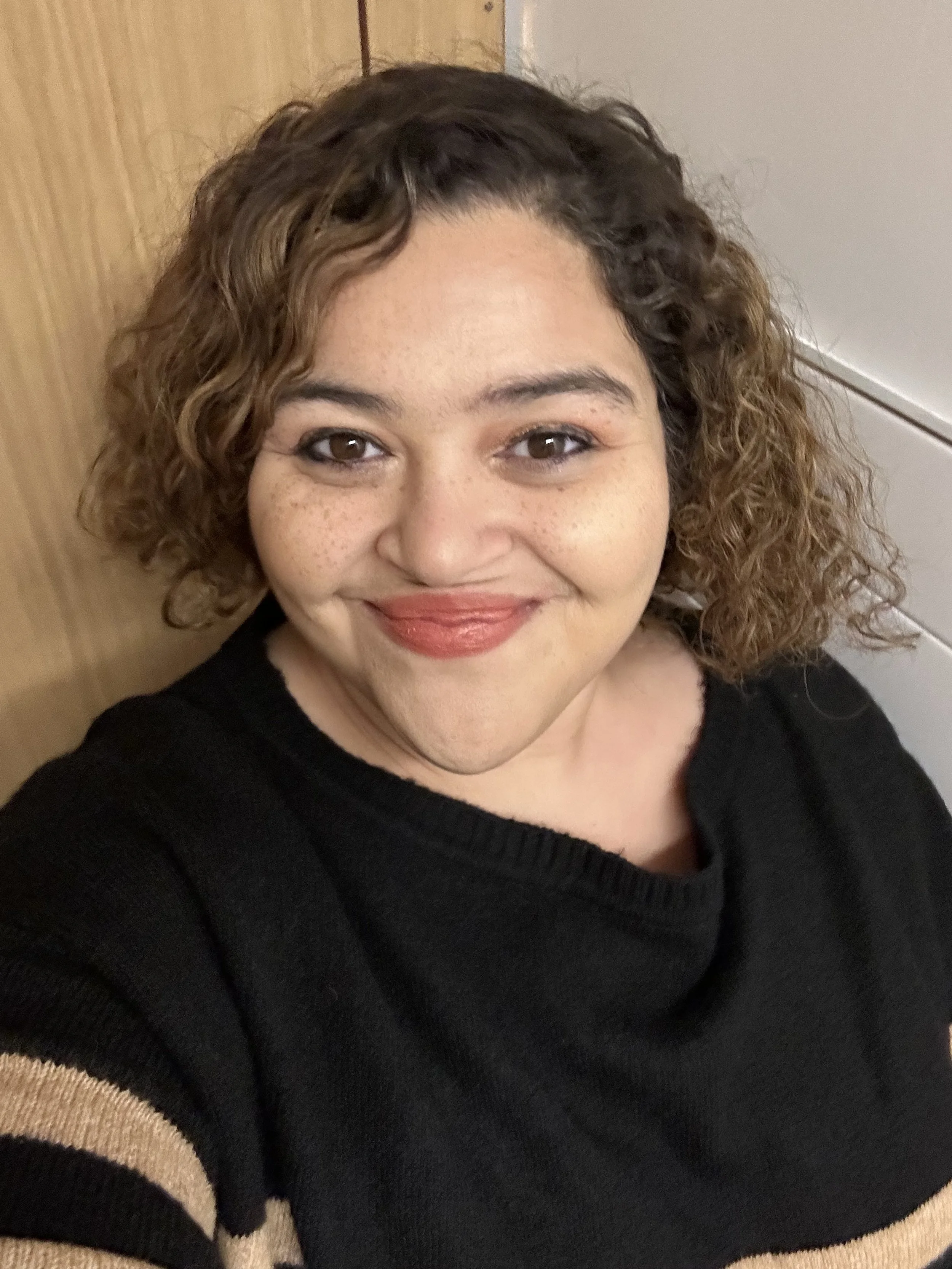 A smiling woman with curly brown hair, wearing a black sweater with tan stripes on the sleeve, taking a selfie indoors against a wooden and white background.