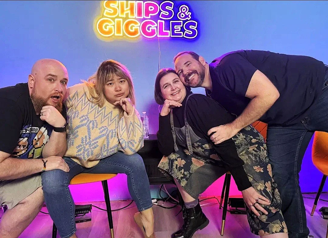 Four people posing together in front of a neon sign that reads 'Ships & Giggles.' They are sitting on orange chairs and smiling, with one person leaning over another. The setting appears to be a comedy or entertainment venue with colorful lighting and a casual atmosphere.