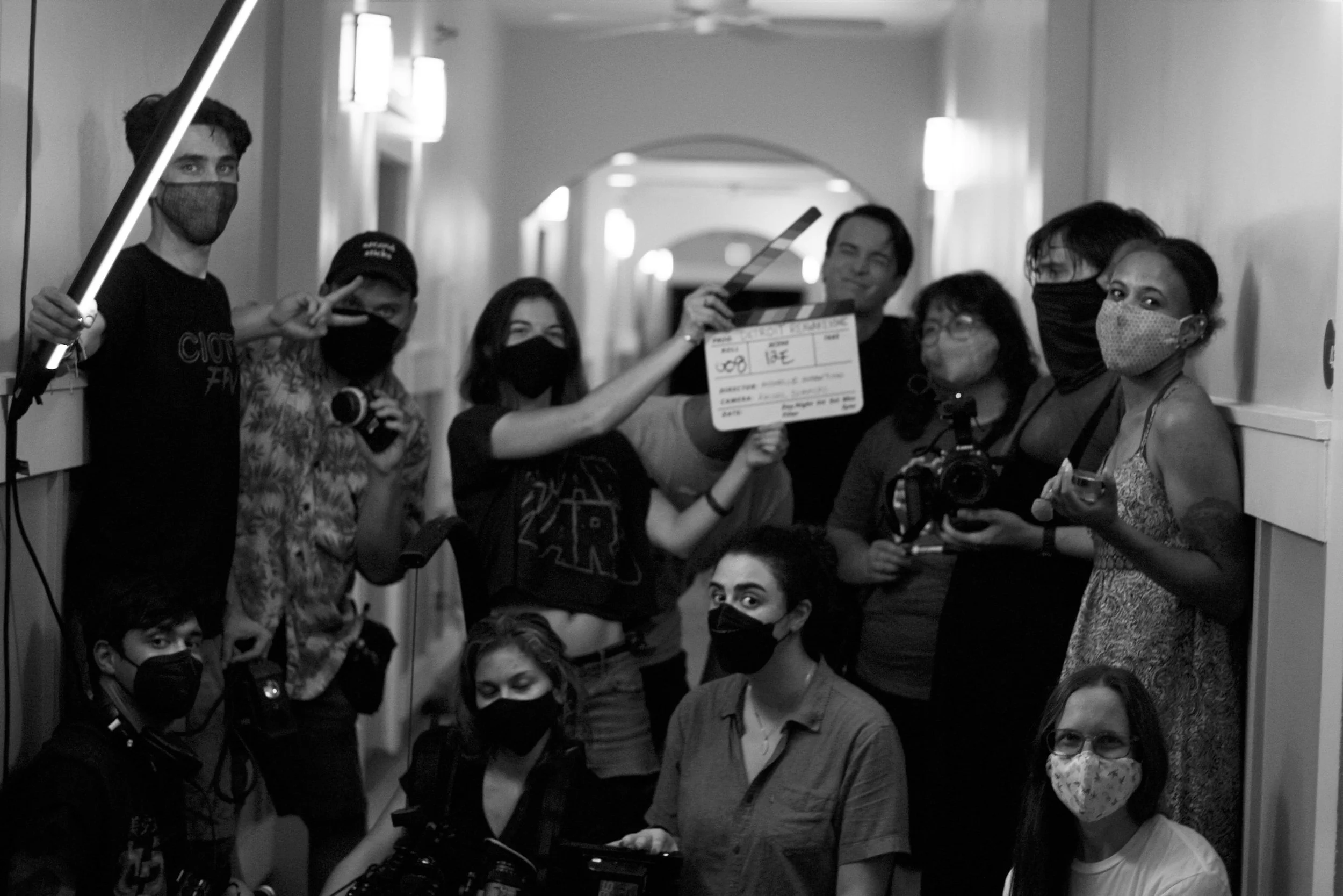 A group of people, all wearing face masks, gathered in a hallway on a film set. They are holding equipment such as cameras, a clapperboard, and a light. Some individuals are posing for the photo, with one person showing a peace sign, suggesting a behind-the-scenes moment during a filming or photography session.