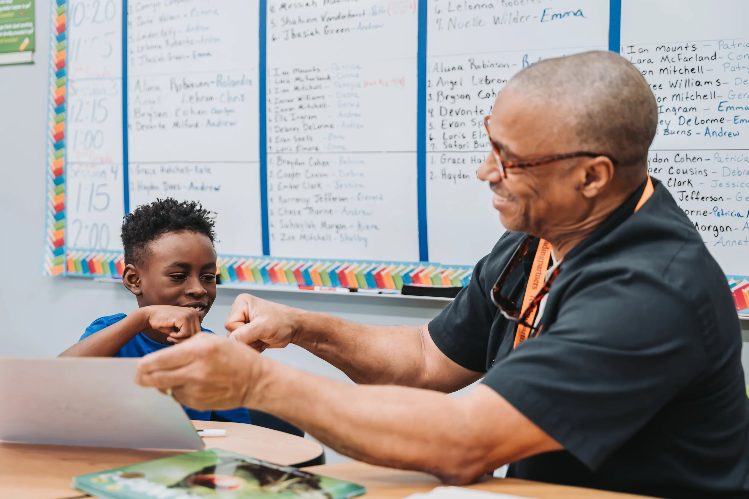 An older elementary student seated at a desk in a classroom giving a high-five to a teacher.
