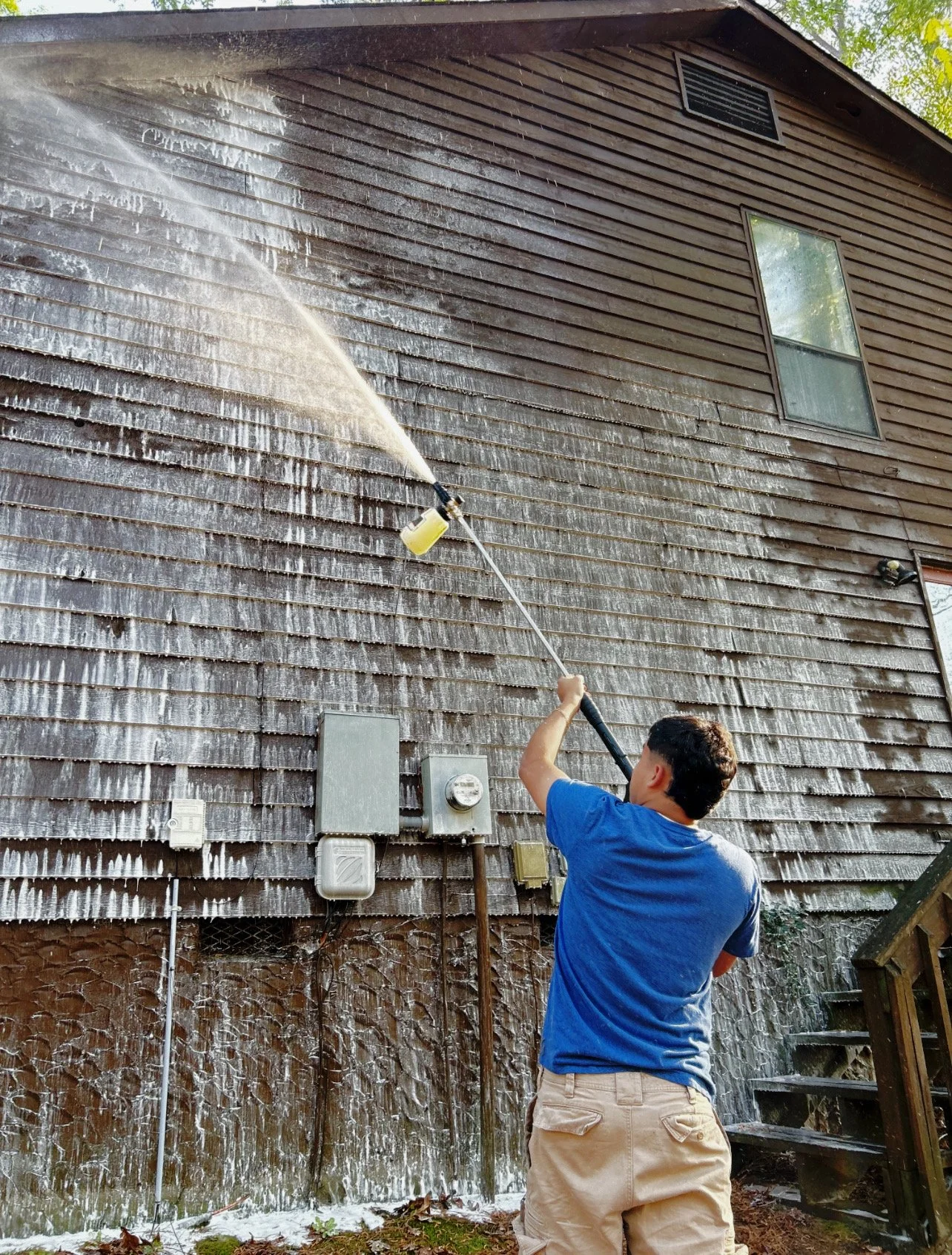 pressure washing the outside wooden siding of a house. The pressure washing creates a large area of cleaned, lighter wood, contrasting with the darker, dirty wood remaining on the house.