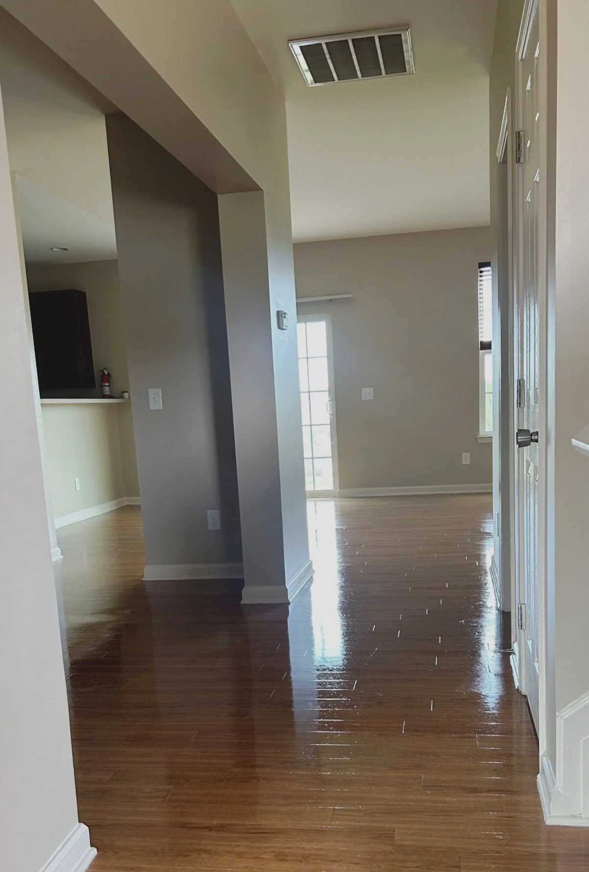 An empty room in a house with wooden floors, beige walls, a sliding glass door, and a window, with parts of a kitchen visible to the left.