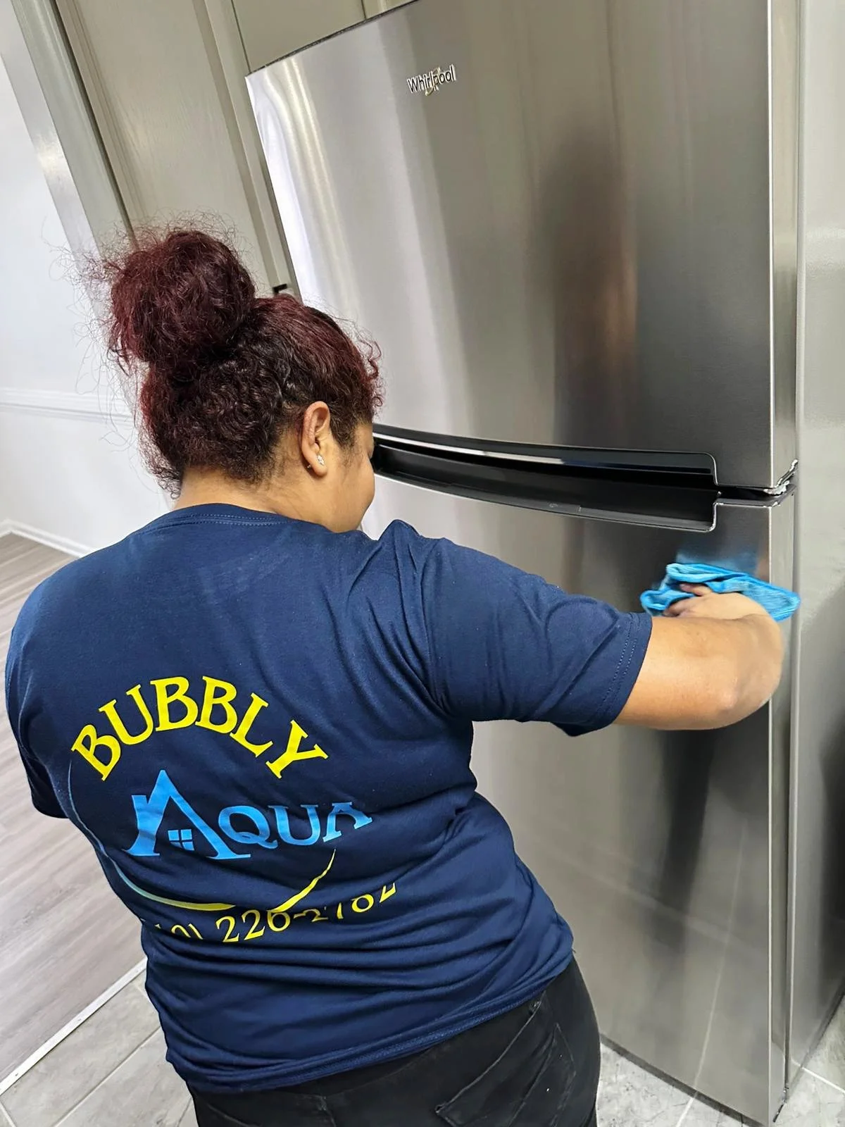 A person cleaning the side of a stainless steel refrigerator with a blue cloth, wearing a navy blue t-shirt that says "BUBBLY AQUA" on the back.