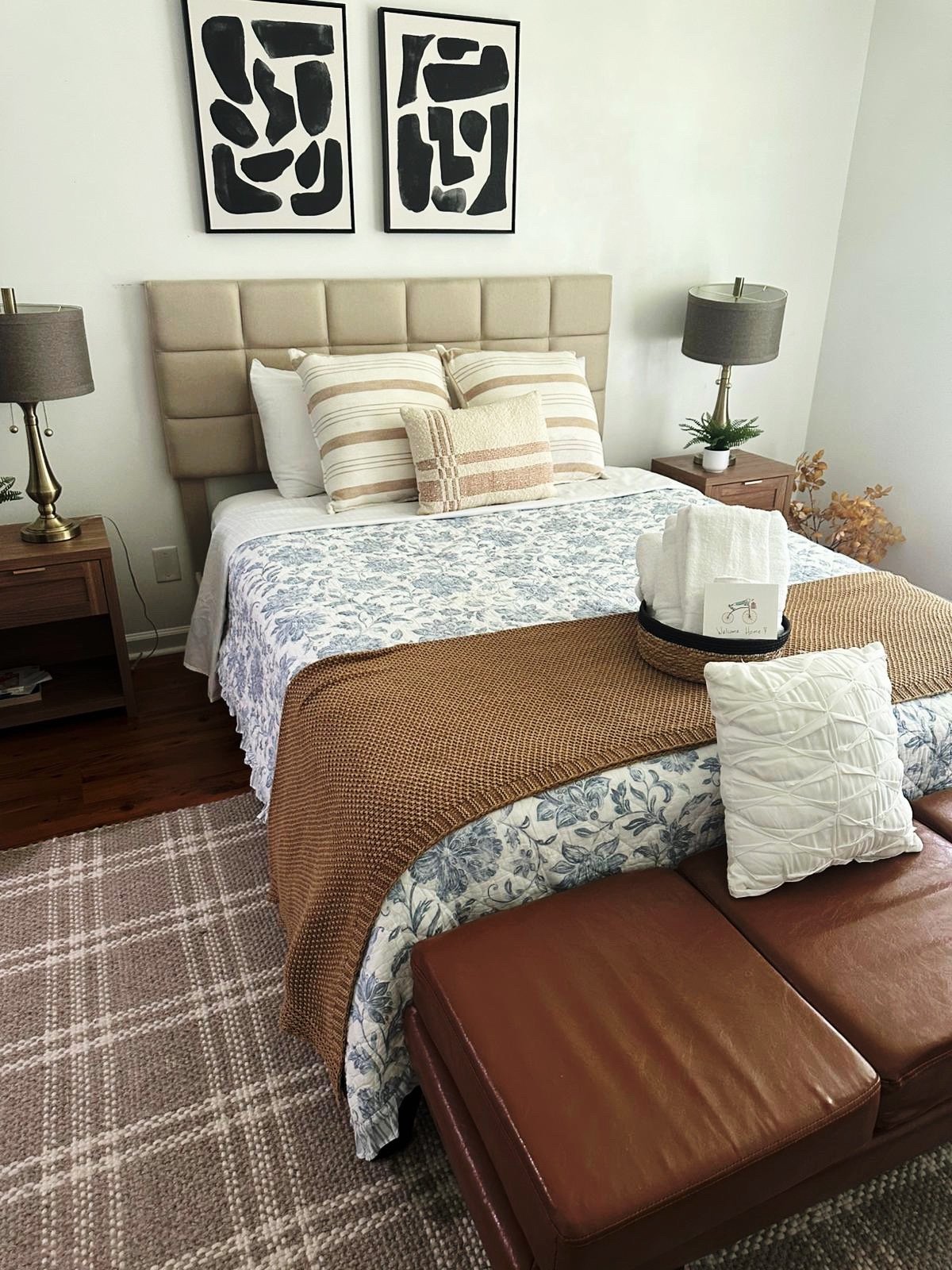 A neatly made bed with beige headboard and striped pillows in a cozy bedroom. Two matching bedside tables with lamps, art on the wall, a basket of towels, and a brown ottoman at the foot of the bed.