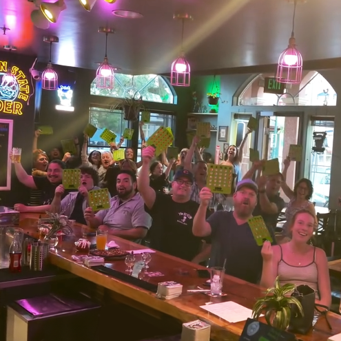 Group of people in a bar celebrating with bingo cards, raising their hands and smiling.