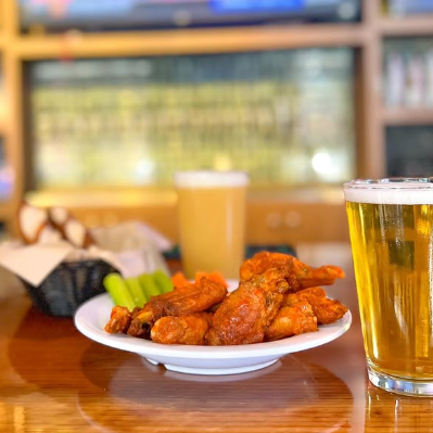 Plate of oven-baked chicken wings with celery and carrot sticks, as well as glasses of beer on a wooden bar.