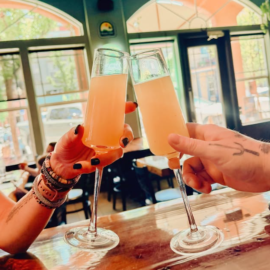 Two people clinking glasses of mimosas inside a comfortably lit bar with large windows.