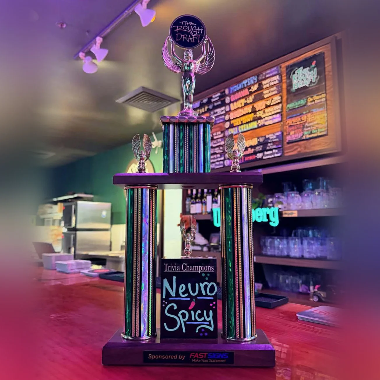 Colorful trophy with a figurine holding a sign that reads 'The Rough Draft', and a chalkboard-style sign with small colorful decorations. The trophy is displayed on a taproom bar counter with a menu board and glasses in the background.