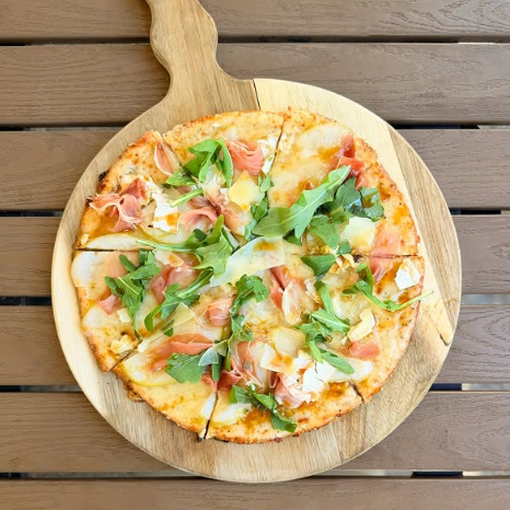 A pizza with brie, parmesan, prosciutto, hot honey, and arugula on a wooden pizza board.