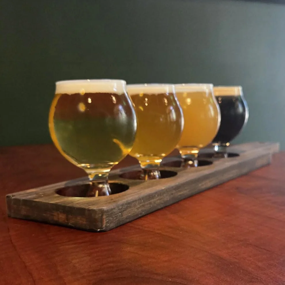 A flight of four different beers in stemmed glasses on a wooden beer flight holder, placed on a wooden table.