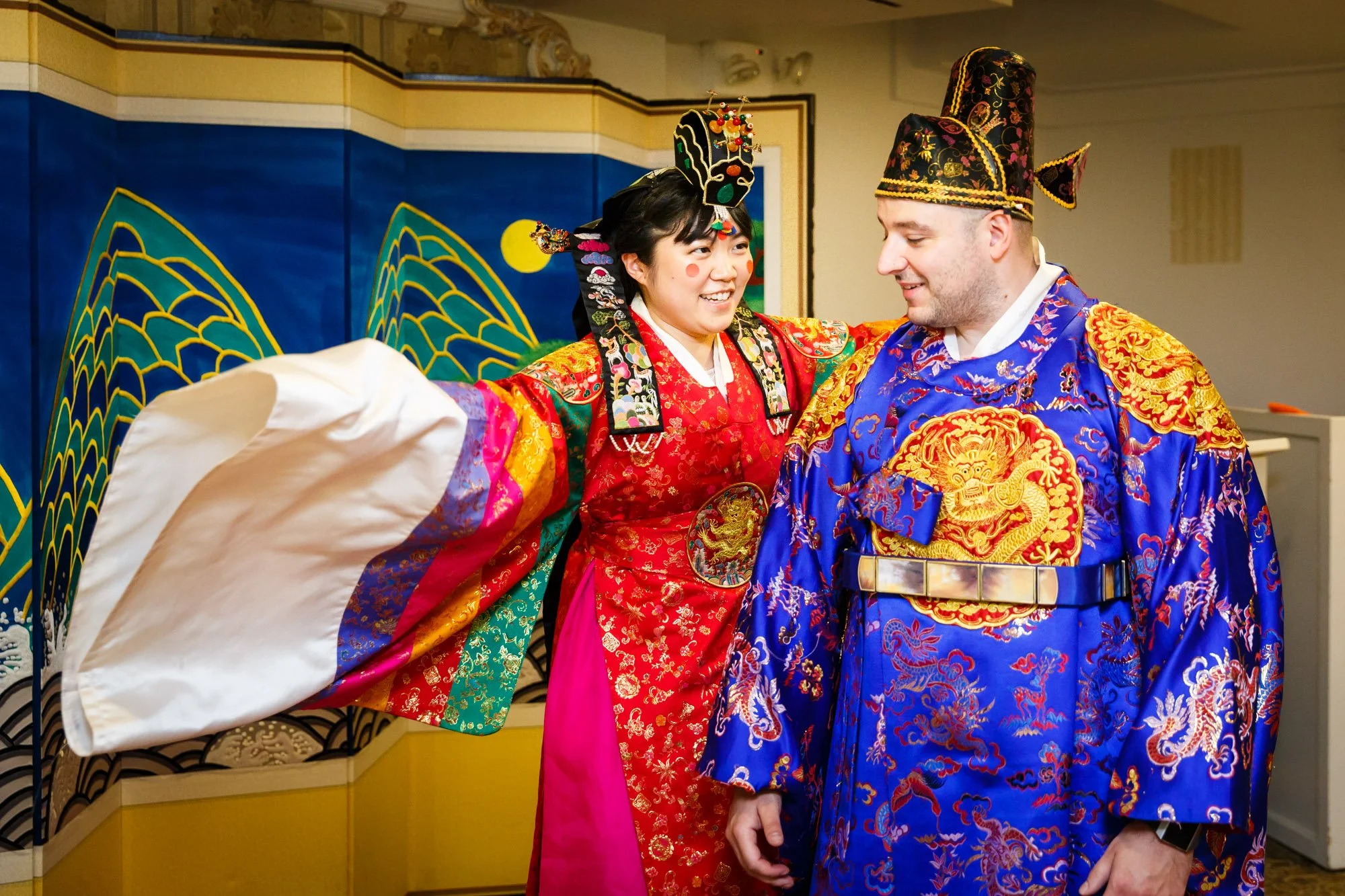 A couple dressed in traditional Asian wedding attire, the woman in a red and black ornate dress with a headdress, and the man in a blue and gold embroidered robe with a tall hat, smiling and facing each other.