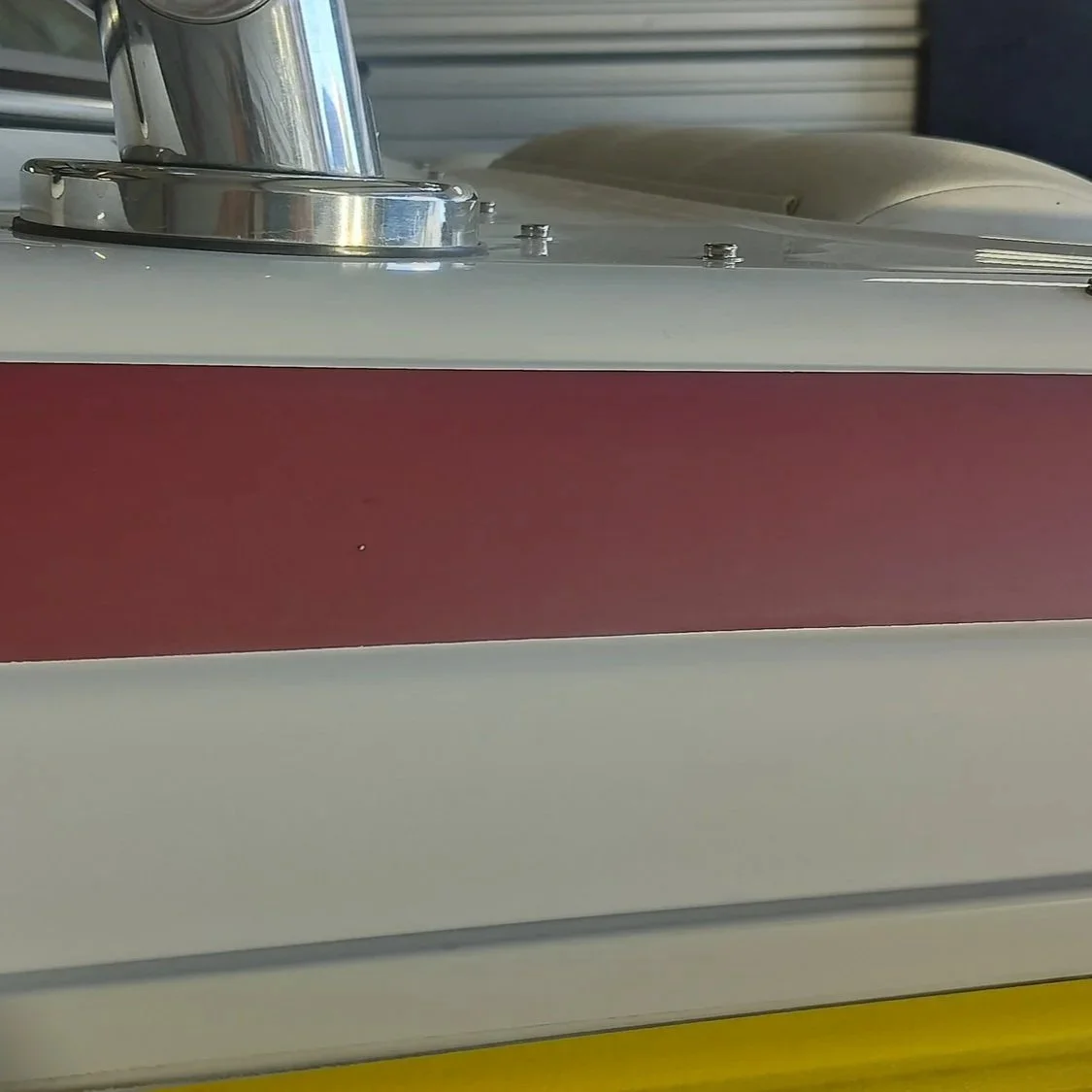 Close-up of a metallic and red and beige machine part, possibly a food processing or manufacturing device, with a shiny metal pipe and screws, set against a background of a metal roll-up door.