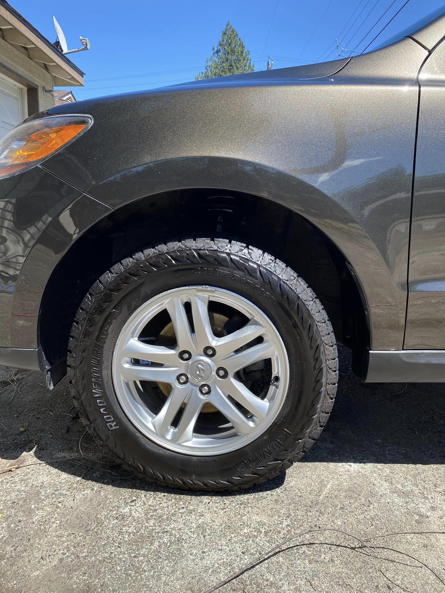 Close-up of a car's front tire and fender, with clear blue sky and a house in the background.
