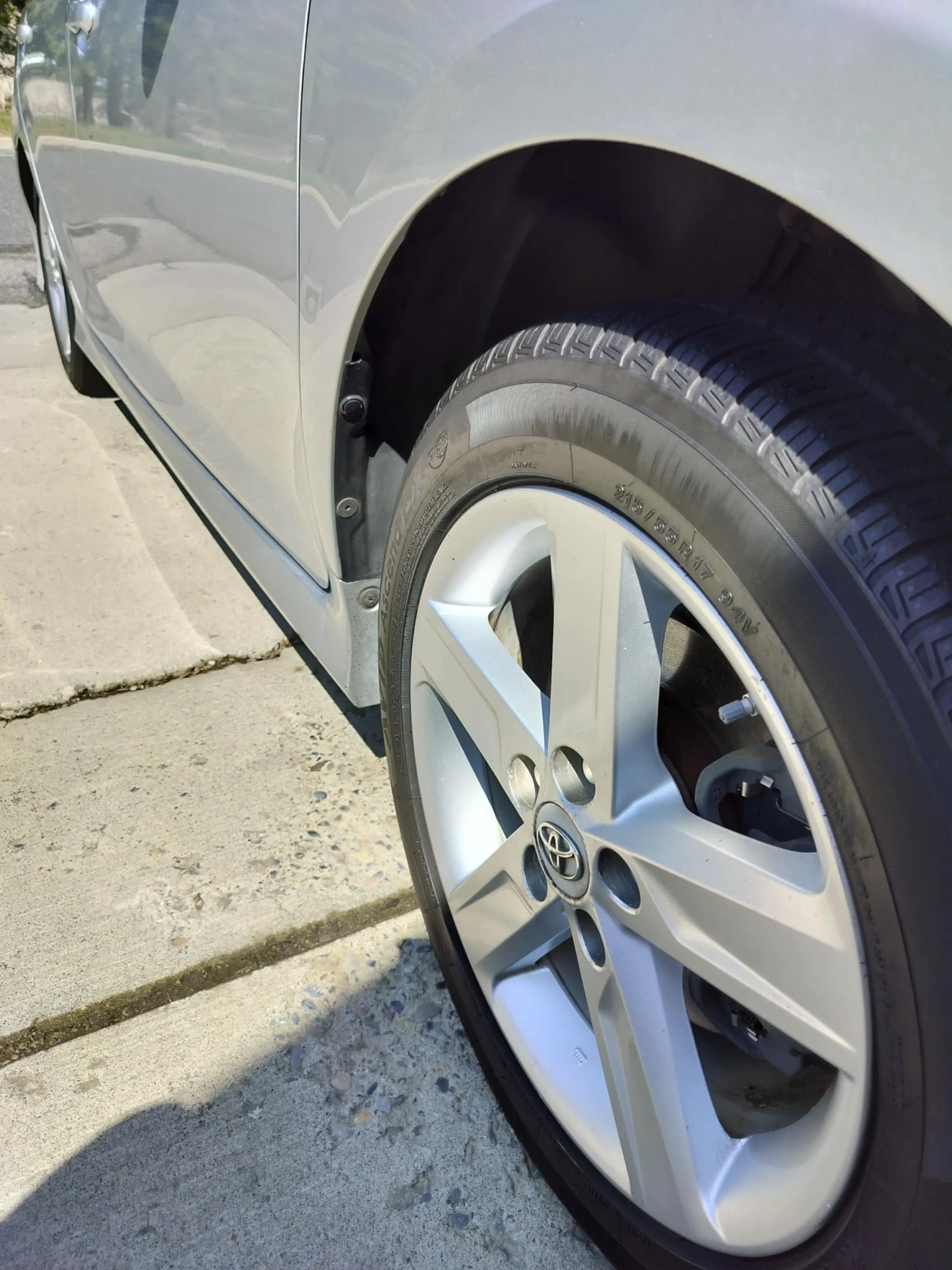 Close-up of a silver Toyota car's front wheel and part of the fender, showing the tire, alloy rim, and vehicle body on a concrete driveway.