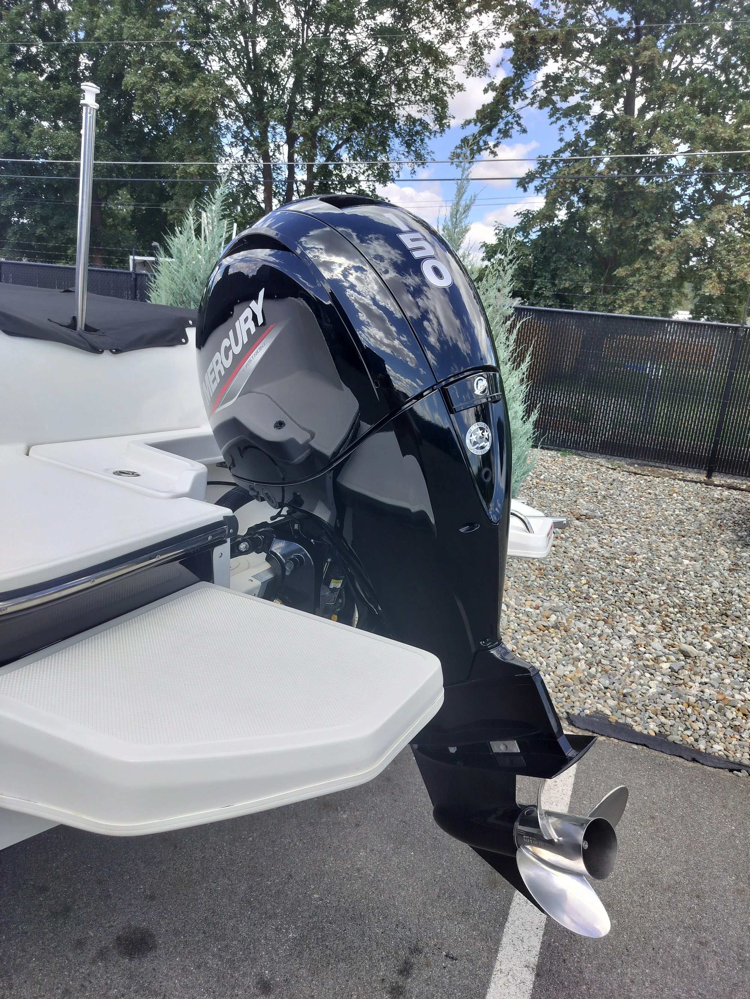 Close-up of a black Mercury outboard motor attached to a boat, with parts of the boat's white hull and a grey platform visible, set outdoors with trees and a black fence in the background.