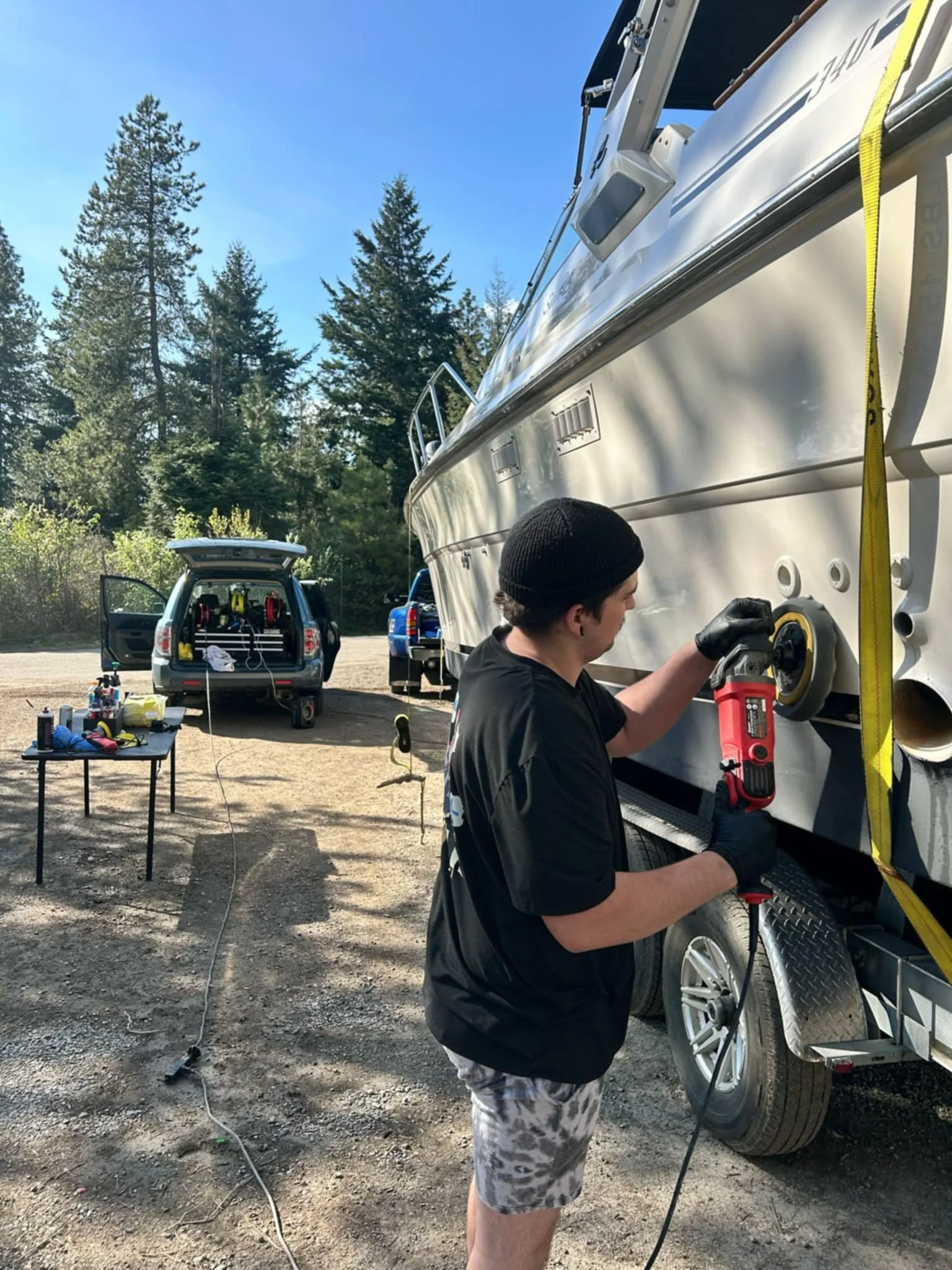 A person wearing a black beanie, black shirt, and tie-dye shorts is polishing or buffing the hull of a boat with a power tool. The boat is on a trailer, and the scene is outdoors with trees and a clear sky in the background. A table with supplies and