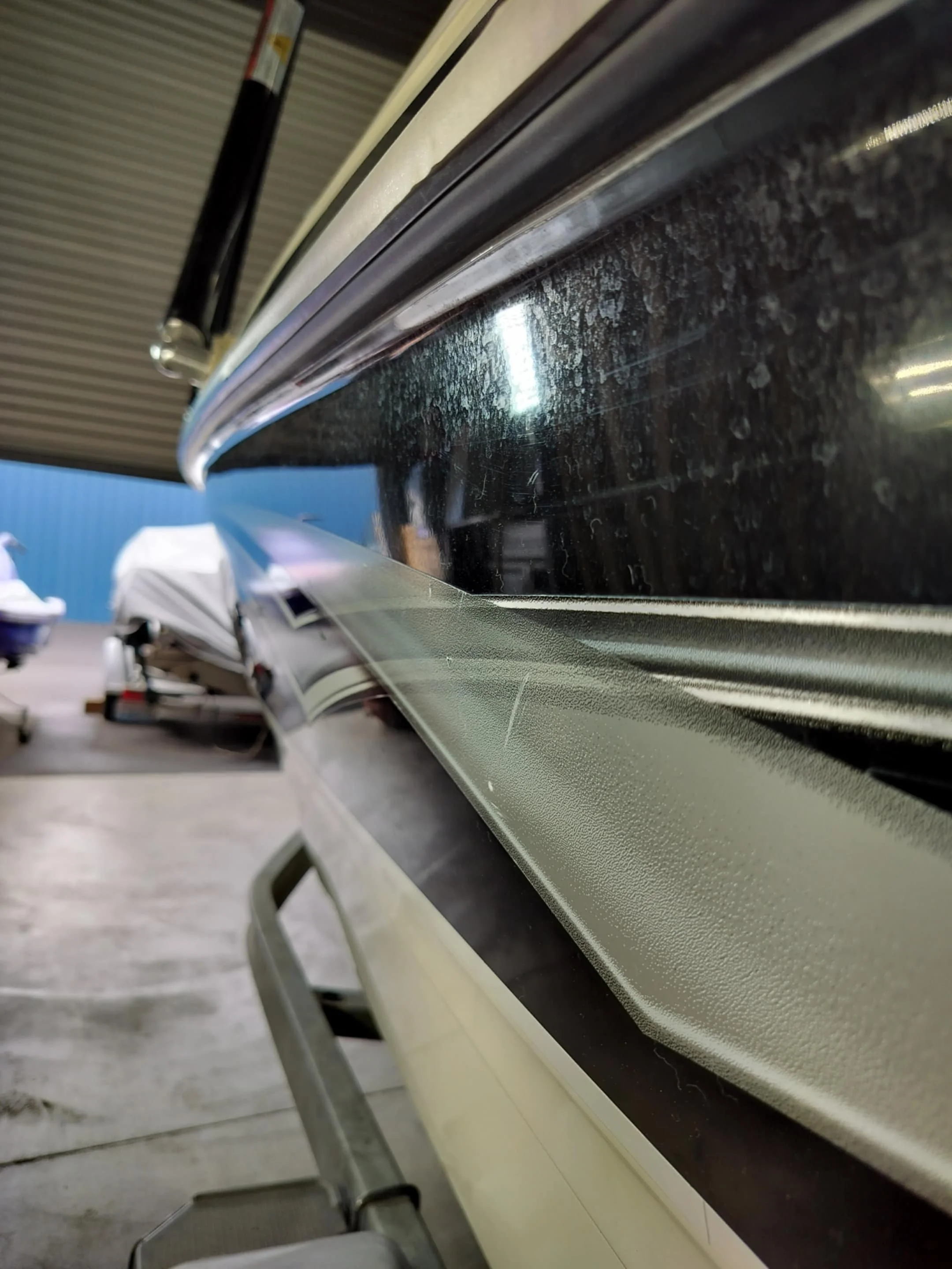 Close-up of the side of a boat in a storage facility, showing the shiny black window and beige textured siding. There is a boat covered with a tarp in the background.
