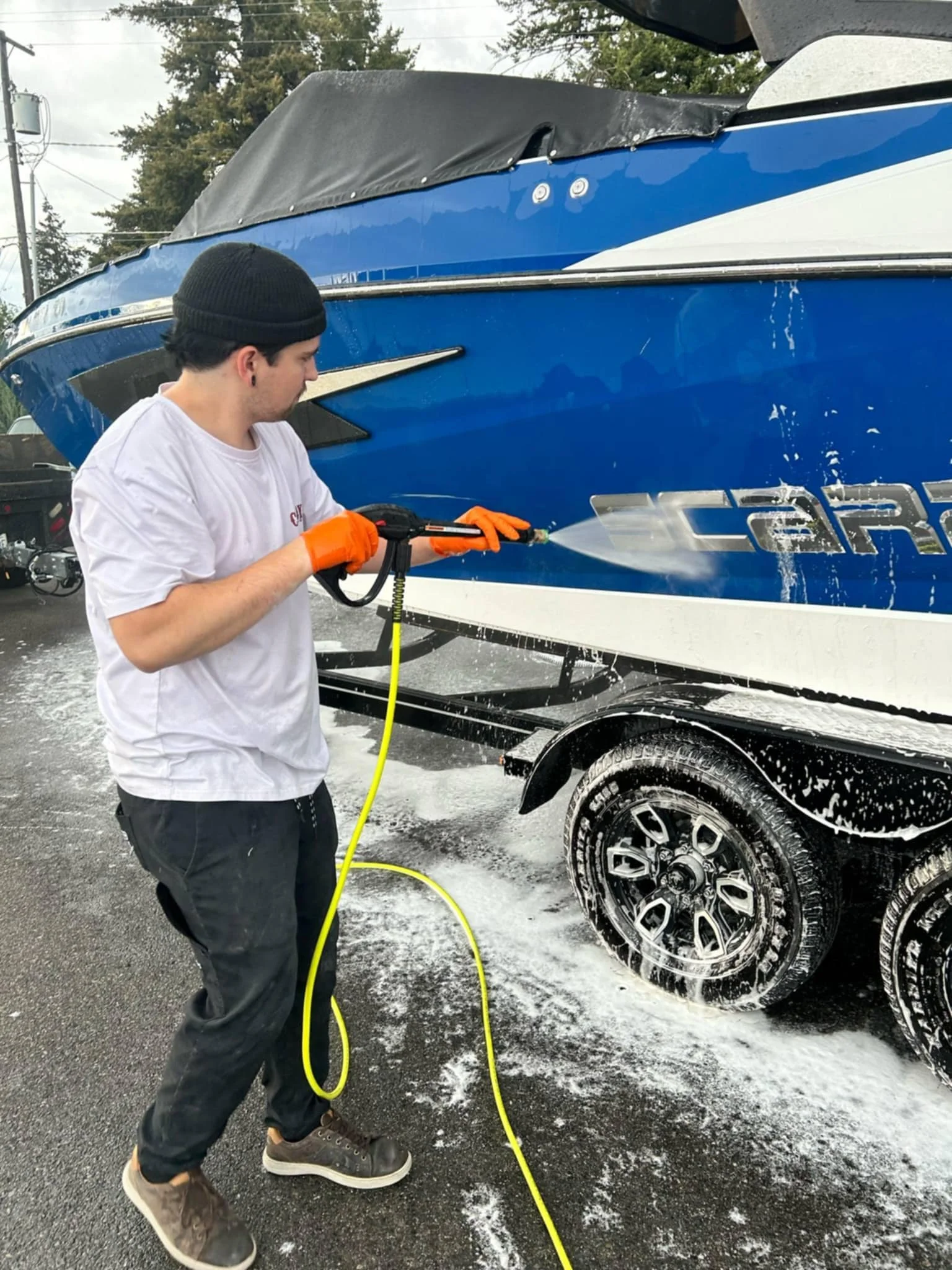 A young man wearing a white t-shirt, black beanie, orange gloves, and black pants is power-washing the side of a blue and white boat on a trailer. Soap and water are visible on the boat and ground.