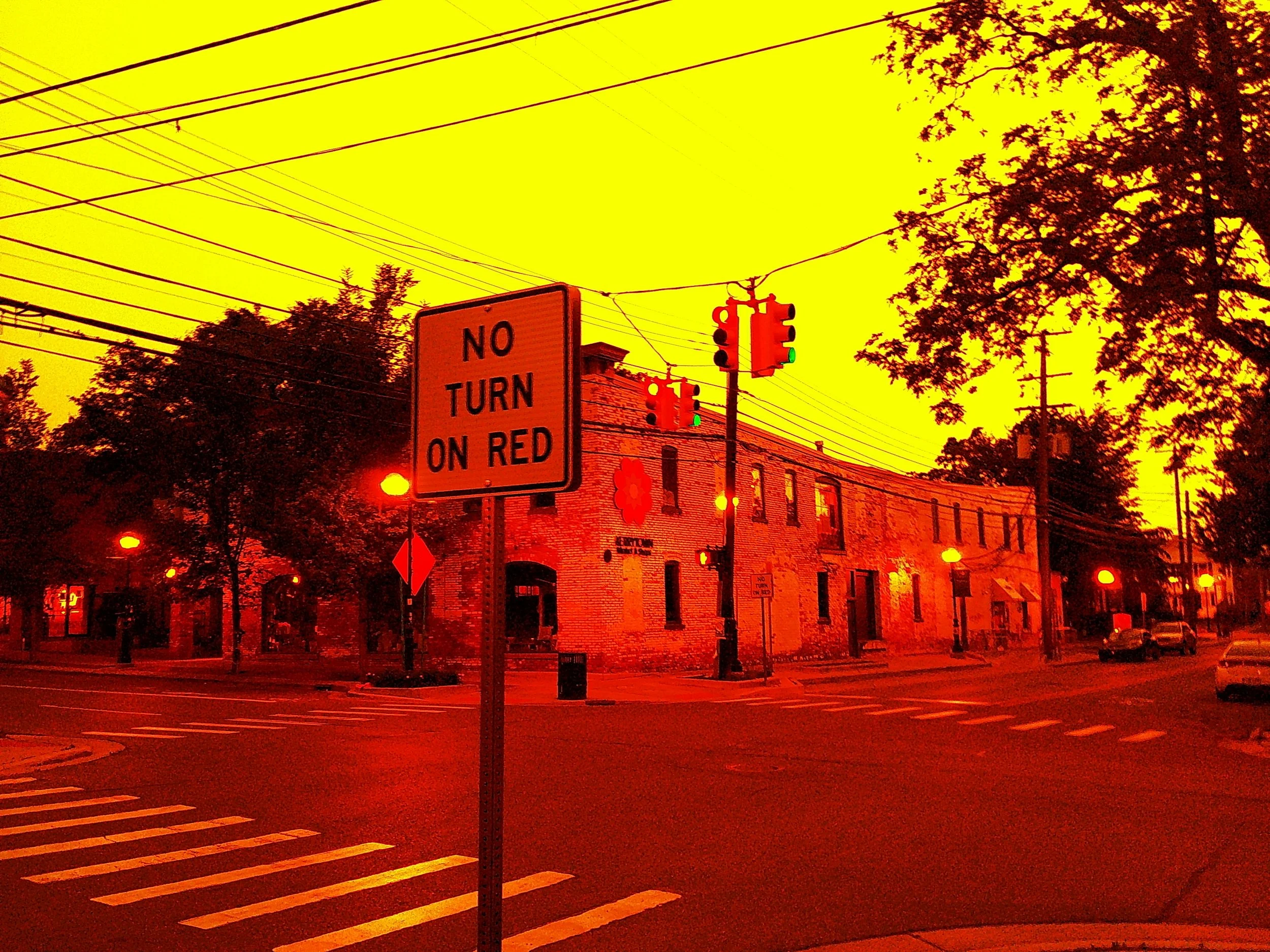 Street scene with traffic lights, a sign that says 'No Turn on Red,' brick buildings, trees, and street lights illuminated with a harsh red and yellow glow.