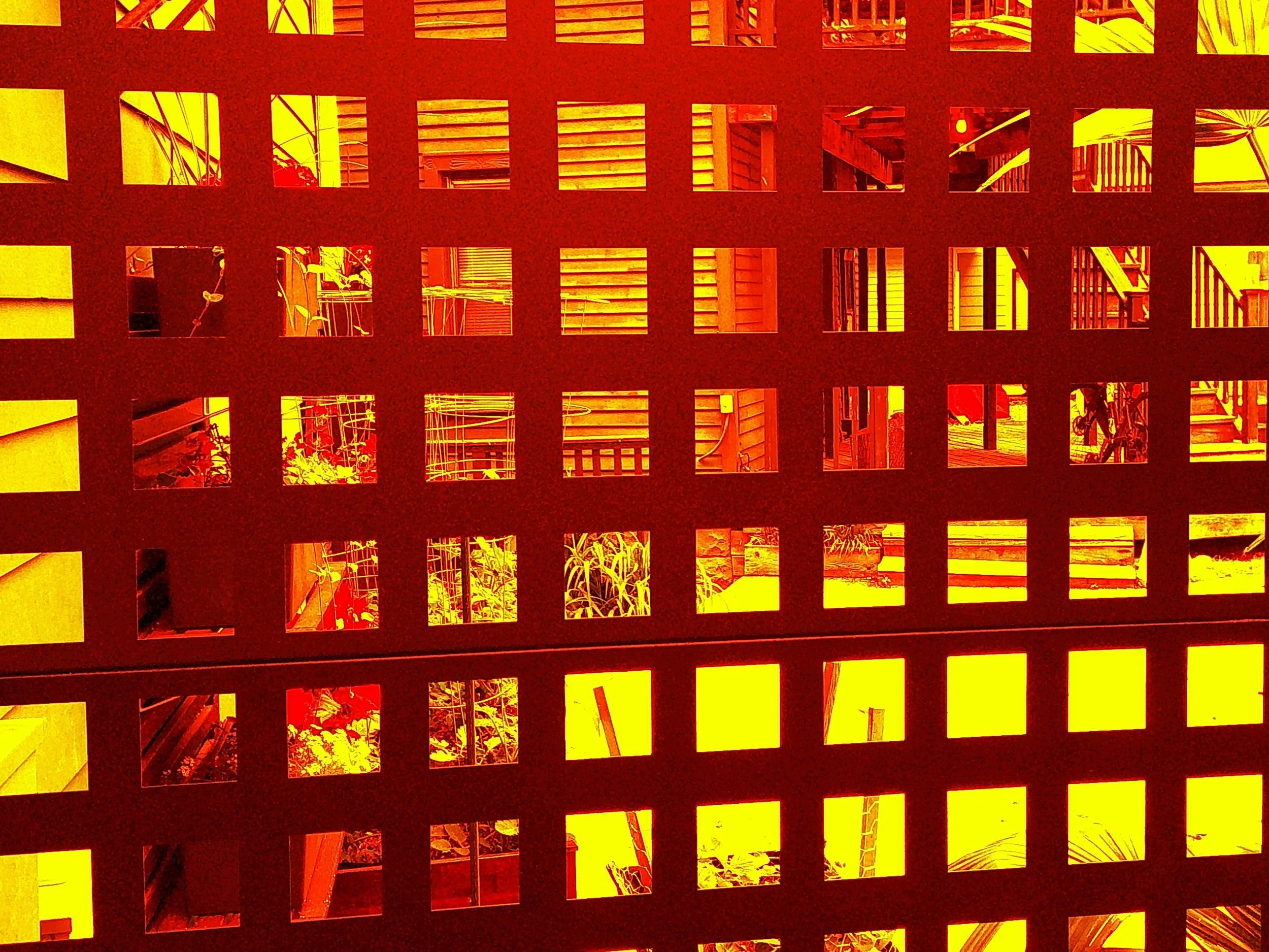 View through a lattice or grid of square openings showing an outdoor area with plants, stairs, and parts of buildings, under warm yellow-orange sunlight.