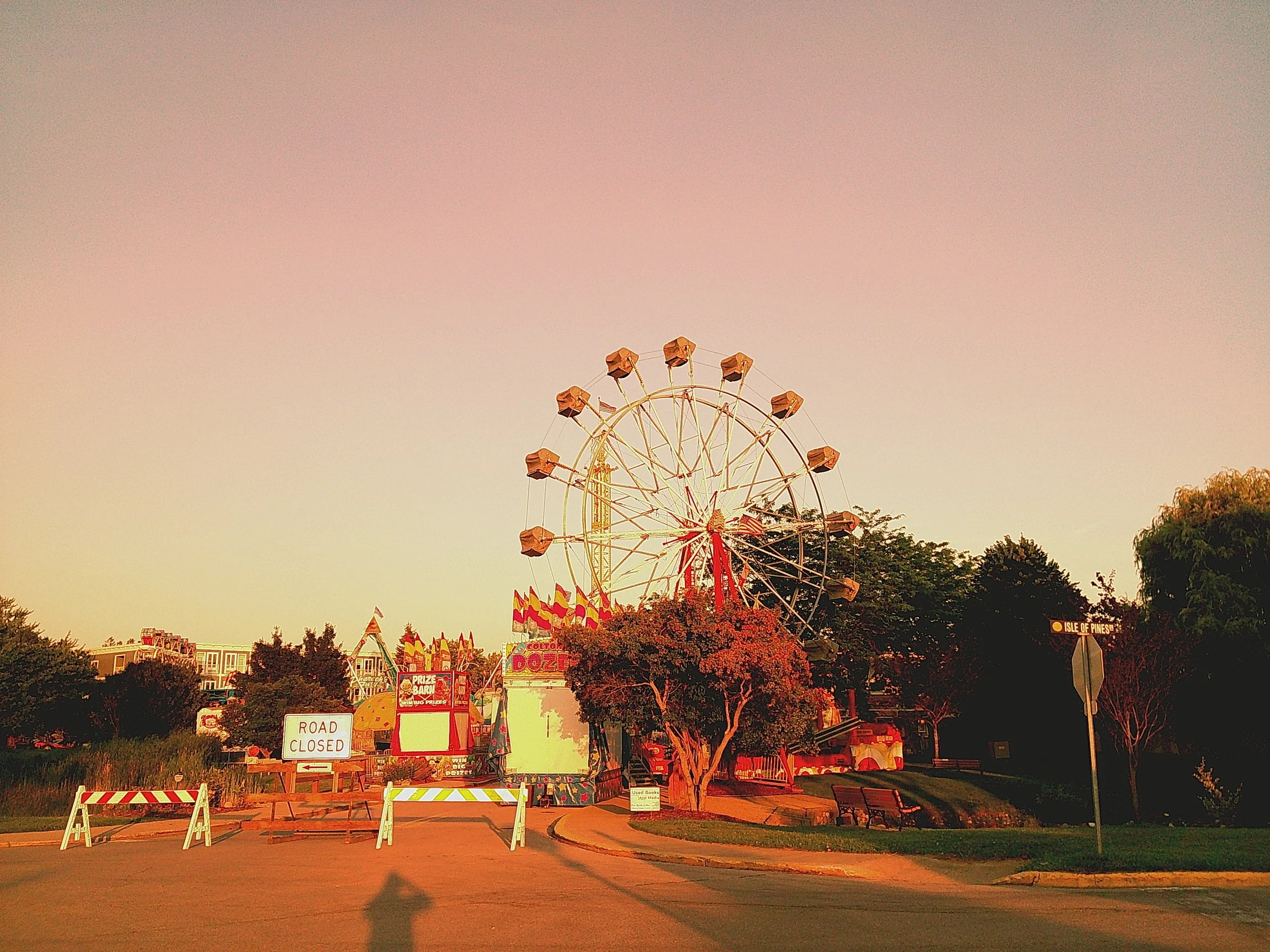 Empty carnival area with a large Ferris wheel, closed road with barriers, and trees at sunset.