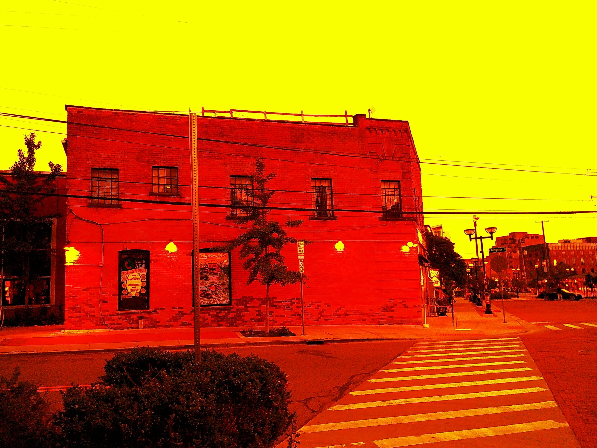 Street view showing a brick building with windows, streetlights, crosswalk, sidewalk, trees, and parked cars, with a yellow sky.