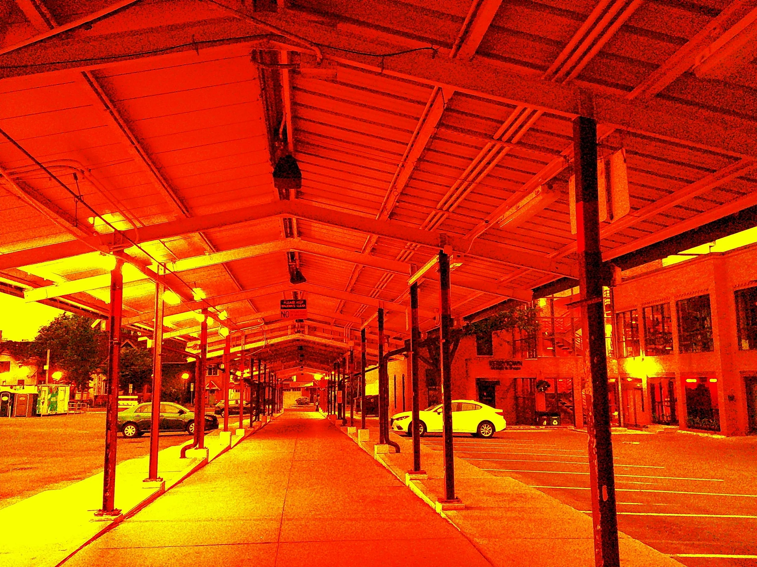 A covered outdoor walkway adjacent to a parking lot with a few parked cars, buildings with large windows, and trees in the background, under a reddish-orange filter.