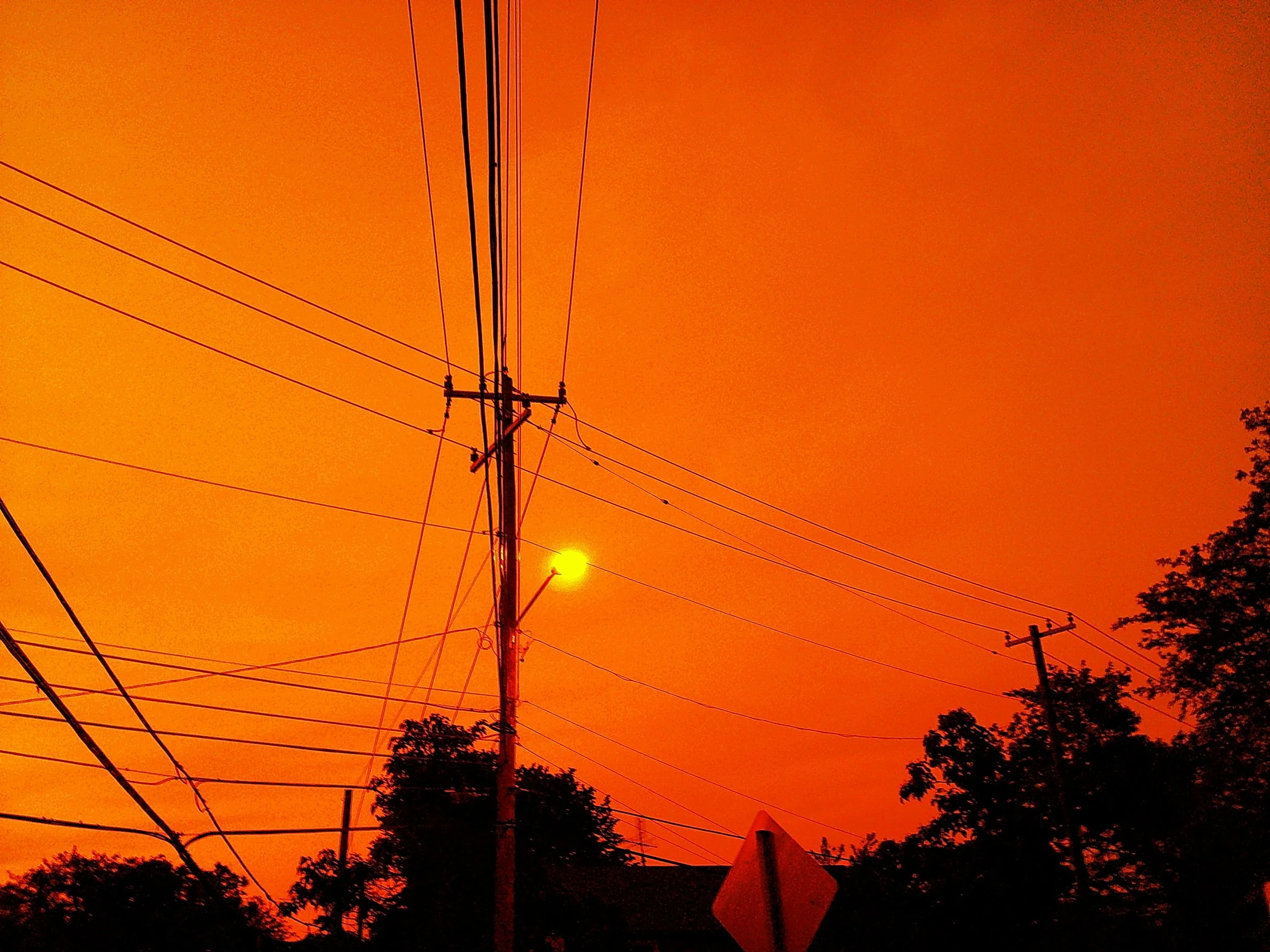 Utility poles and power lines silhouetted against a vivid orange-red sunset or sunrise sky, with trees in the foreground.