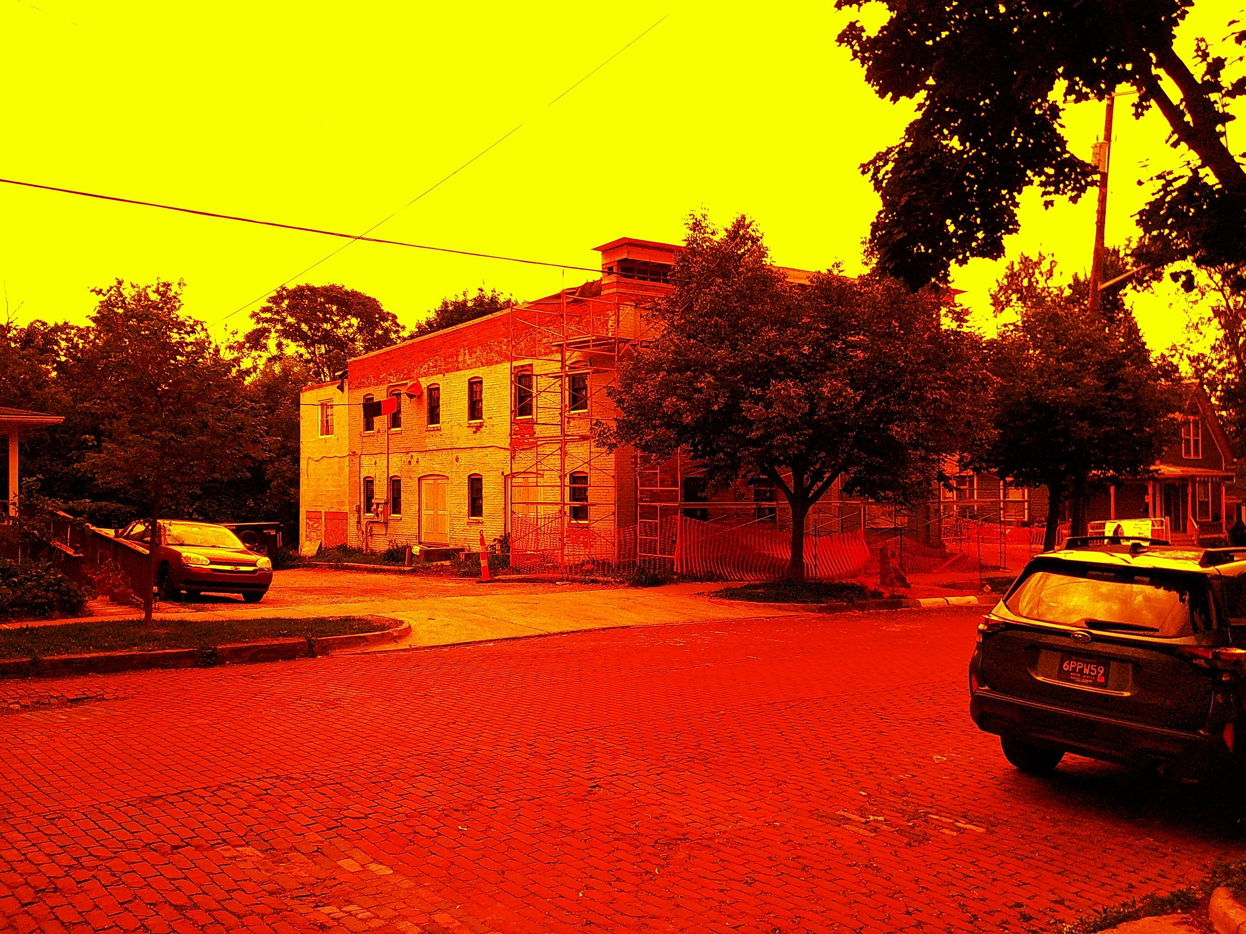 Street view of a brick sidewalk and street with parked cars, trees, and a building under construction, with a yellow sky.