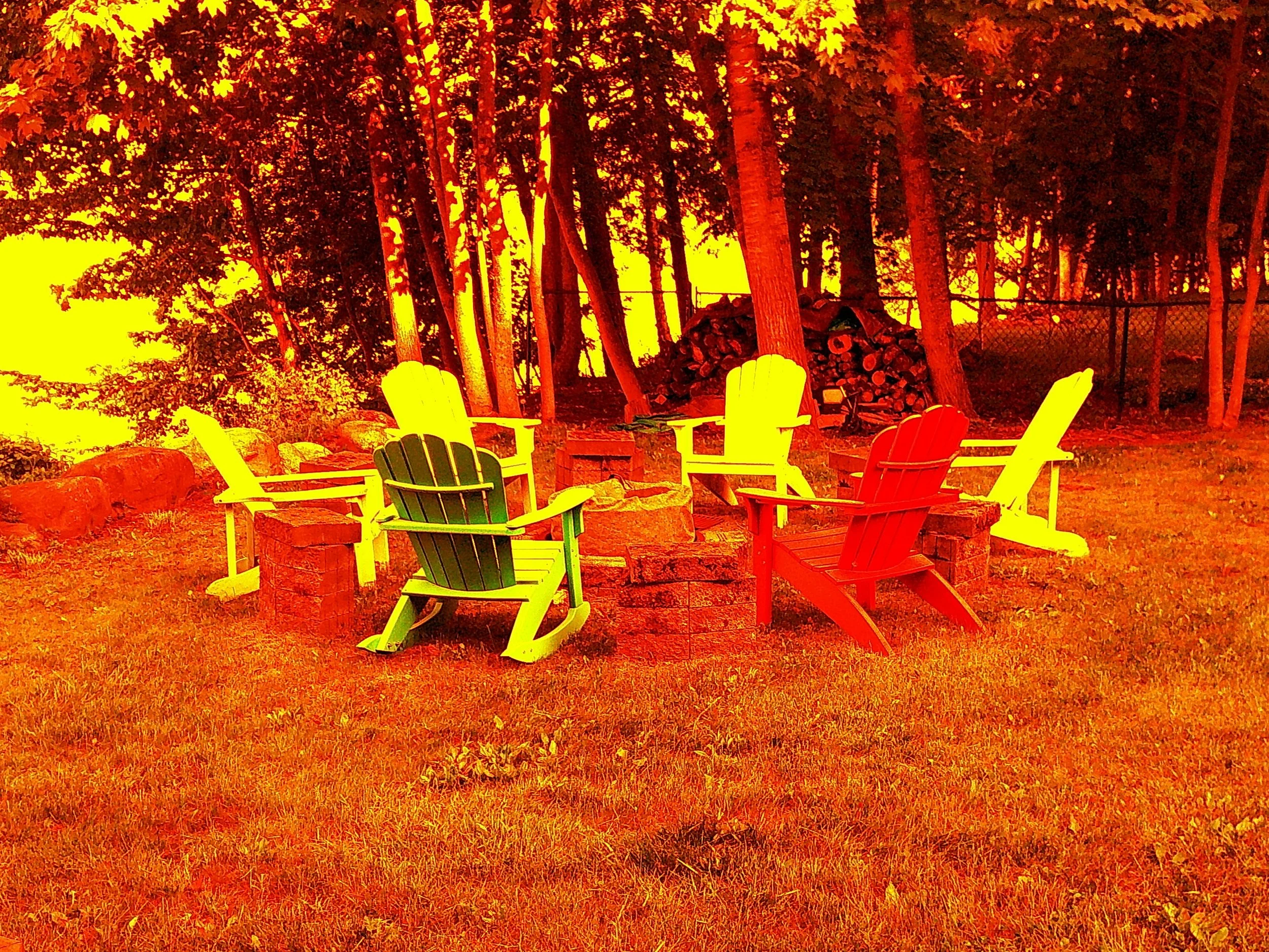 An outdoor scene featuring a circle of colorful Adirondack chairs arranged around a fire pit, set against a background of trees and a woodpile.